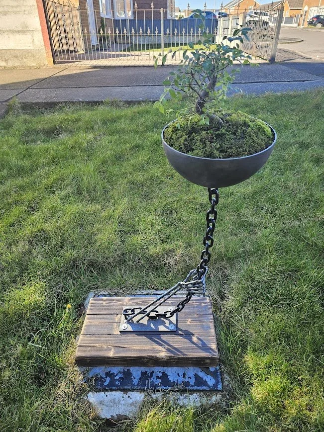 A decorative outdoor stand with a planter on top containing a small tree or shrub, attached to a wooden base with metal chains.