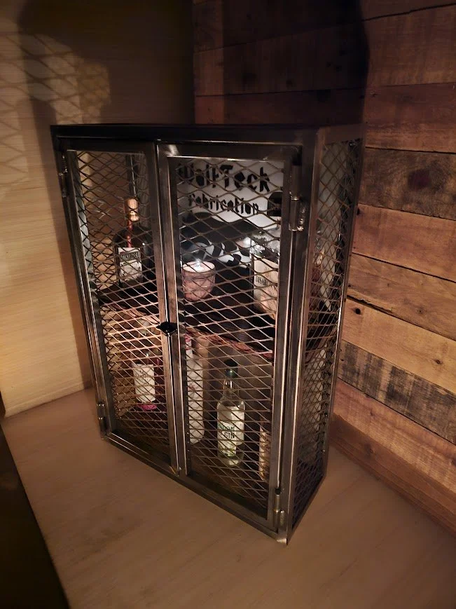 A black metal liquor cabinet with wire mesh doors holding bottles of alcohol inside, placed against a wooden wall.