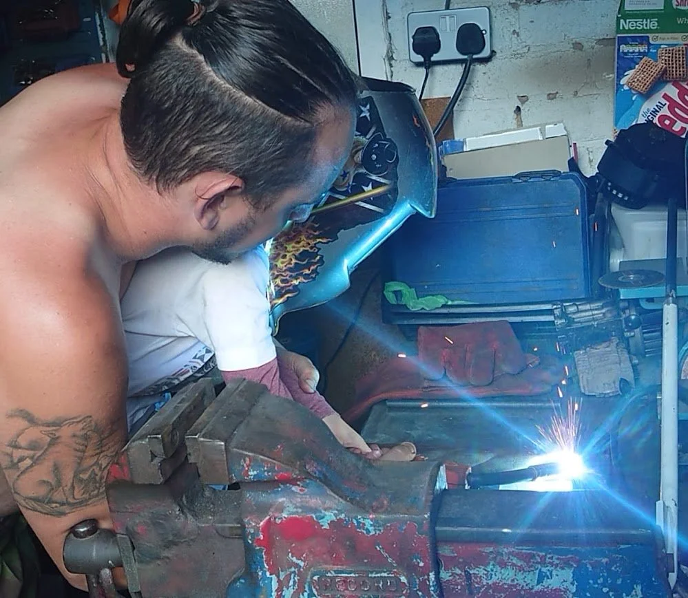 A man is welding metal at a workbench in a garage or workshop, with sparks flying from the welding process.