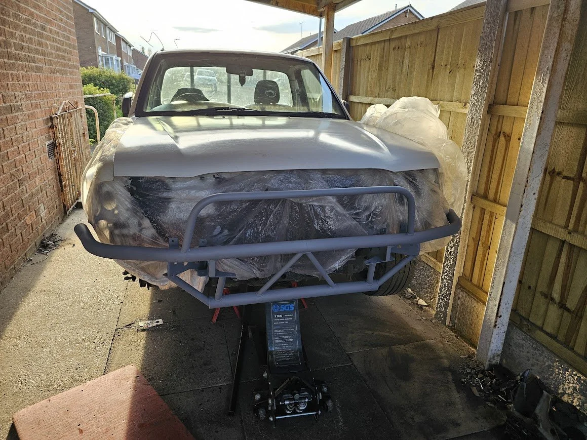 A car being restored or painted, covered with protective plastic, on a car jack in a driveway with a wooden fence on the right and a brick house on the left.