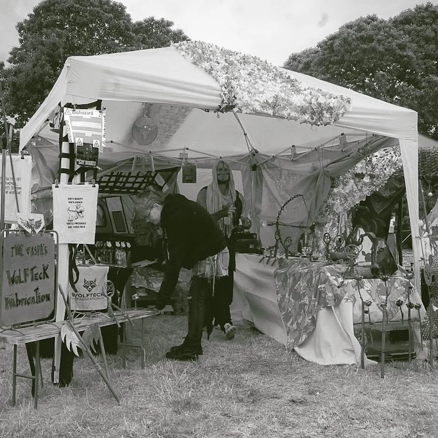 A black and white photo of a vendor booth at an outdoor event, decorated with flowers and fabric, selling jewelry and art pieces. Two people are browsing the items.