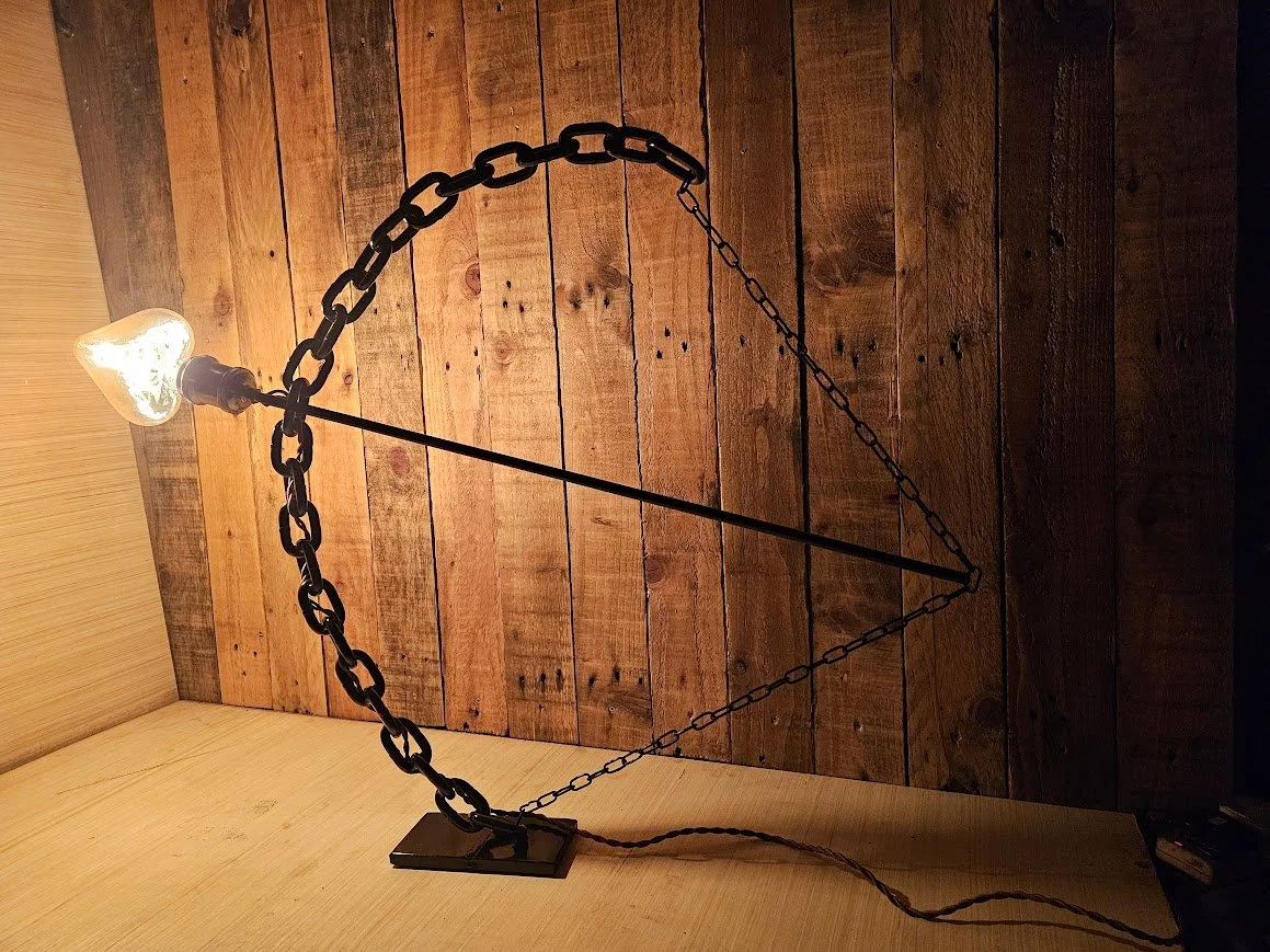 A decorative table lamp with a light bulb, a oranged patterned glass shade, and black chain design, placed on a wooden surface against a wooden plank wall background.