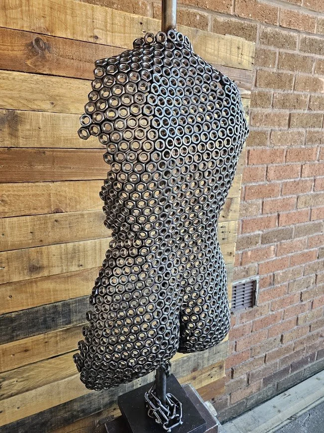 Sculpture of a female torso made from stacked soda cans, mounted on a stand against a wood-paneled wall and brick wall background.