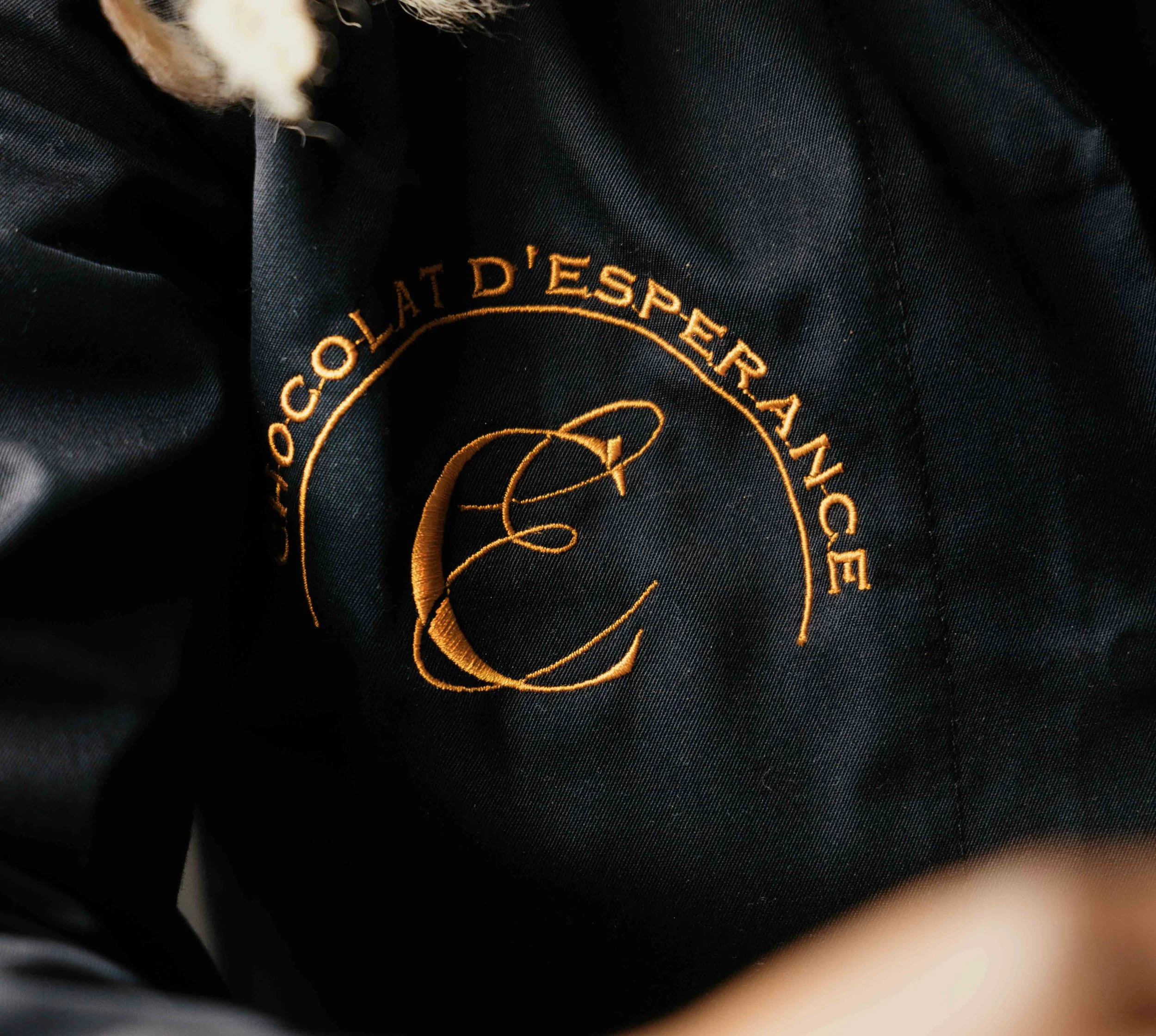 Zoom sur une étiquette brodée en fil doré sur un tissu noir, avec le texte 'Chocolat d'Explorerance' et un symbole graphique en forme de 'E' stylisée.