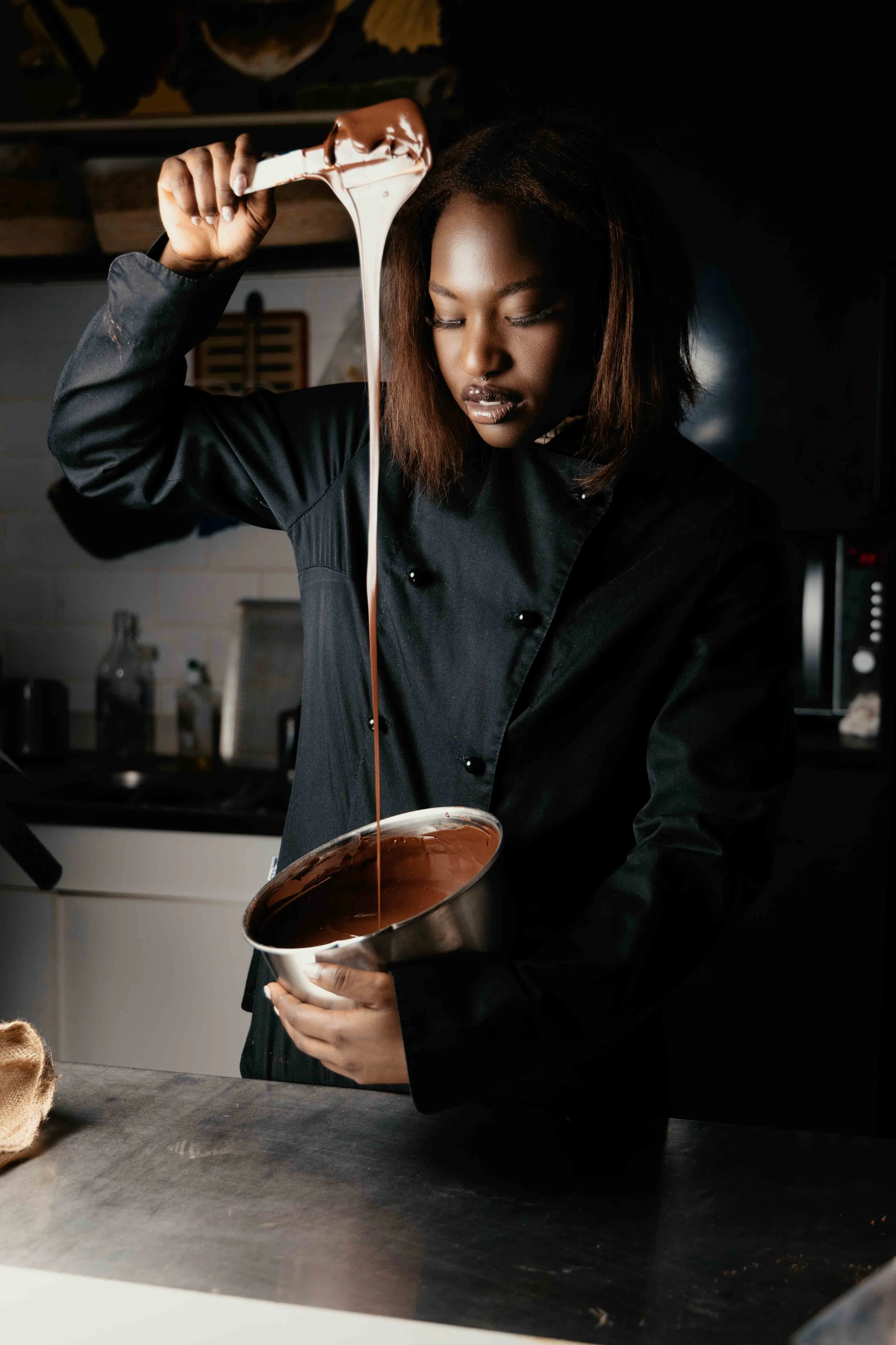 Une femme en cuisine prépare du chocolat fondu dans un bol en acier, devant un plan de travail.