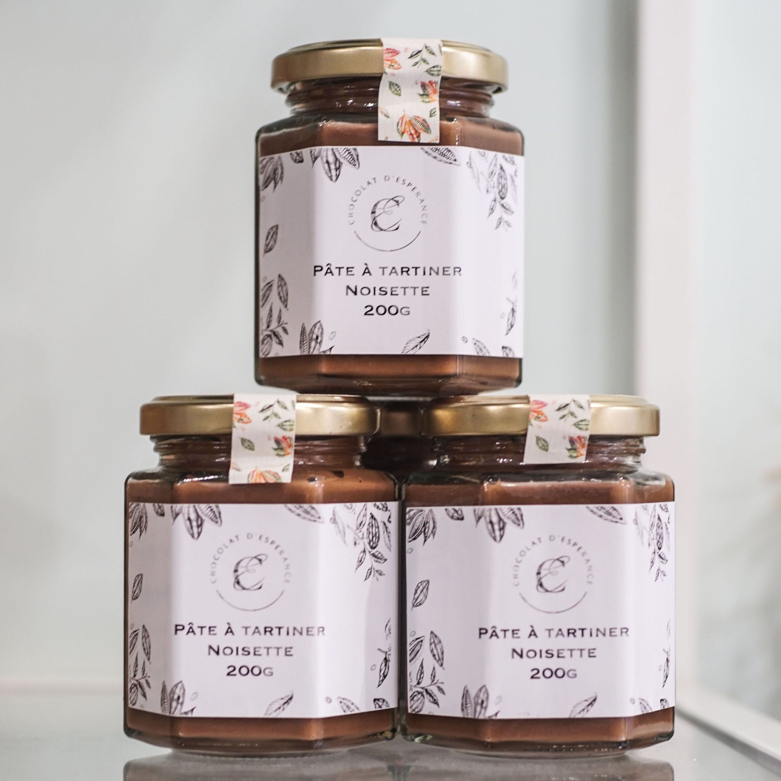 Trois petits pots de pâte à tartiner noisette empilés. Leurs étiquettes indiquent 200g, avec un design floral noir et blanc.