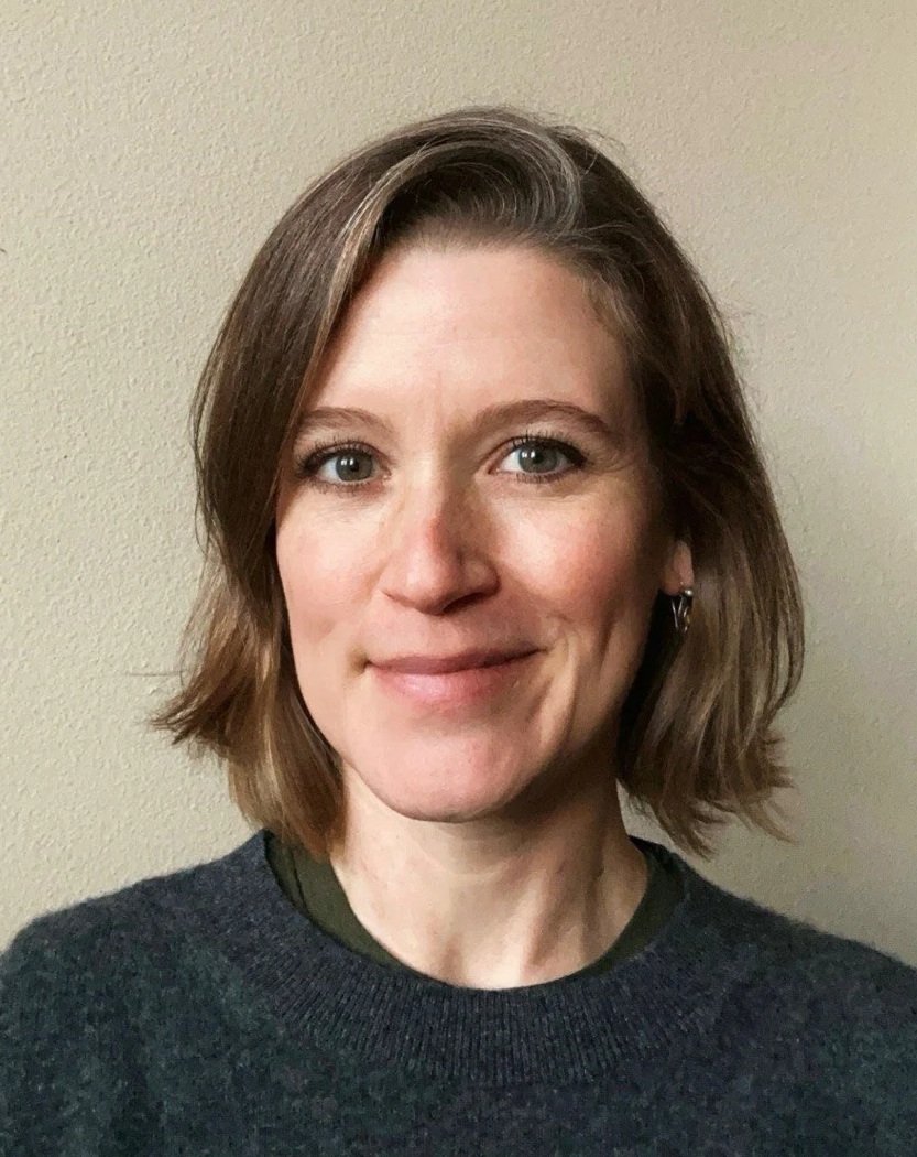A woman with shoulder-length brown hair, blue eyes, and a slight smile, wearing a dark sweater, standing against a beige wall.
