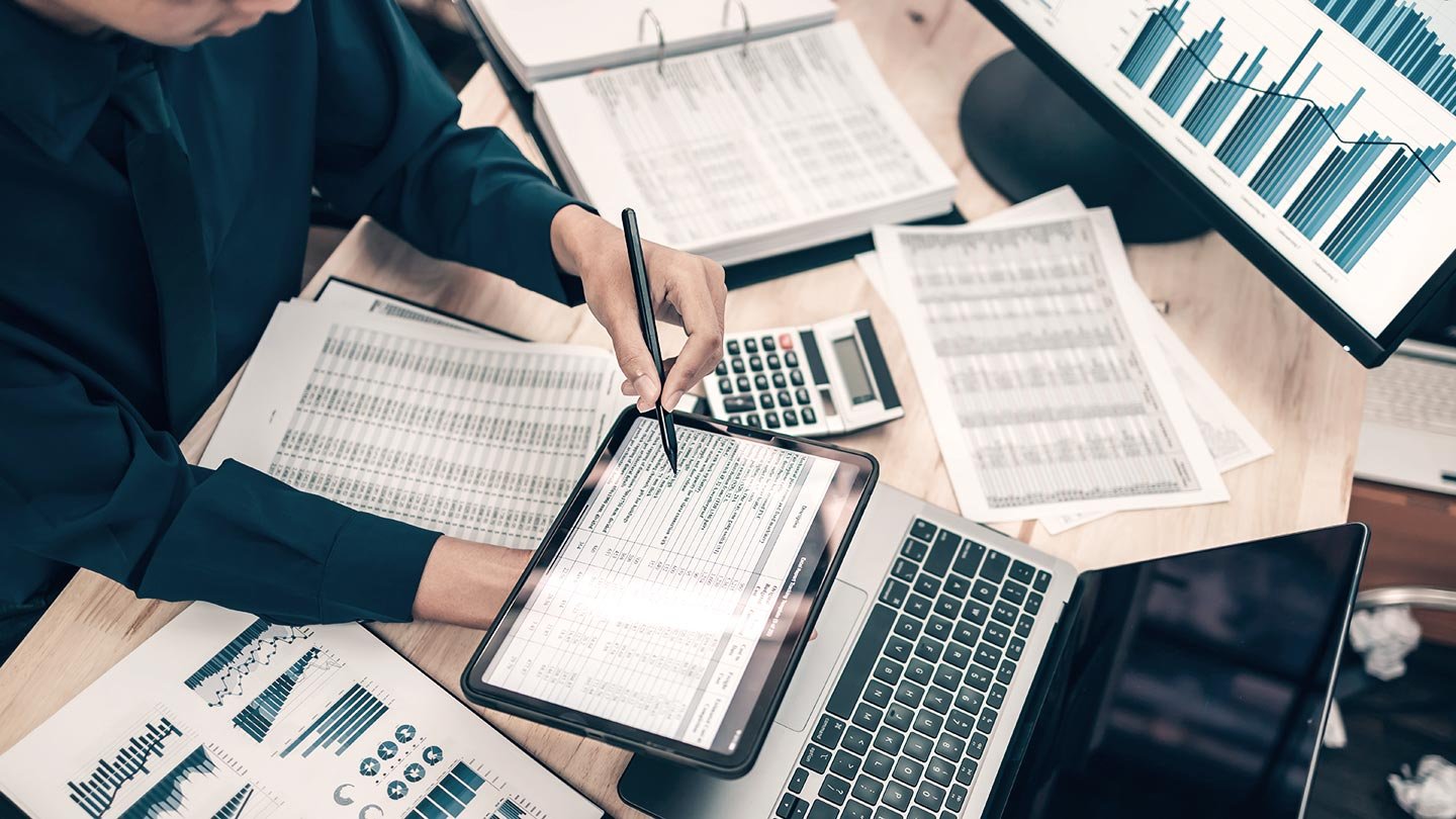 Person working at a desk with financial documents, calculator, laptop, and a tablet displaying data charts