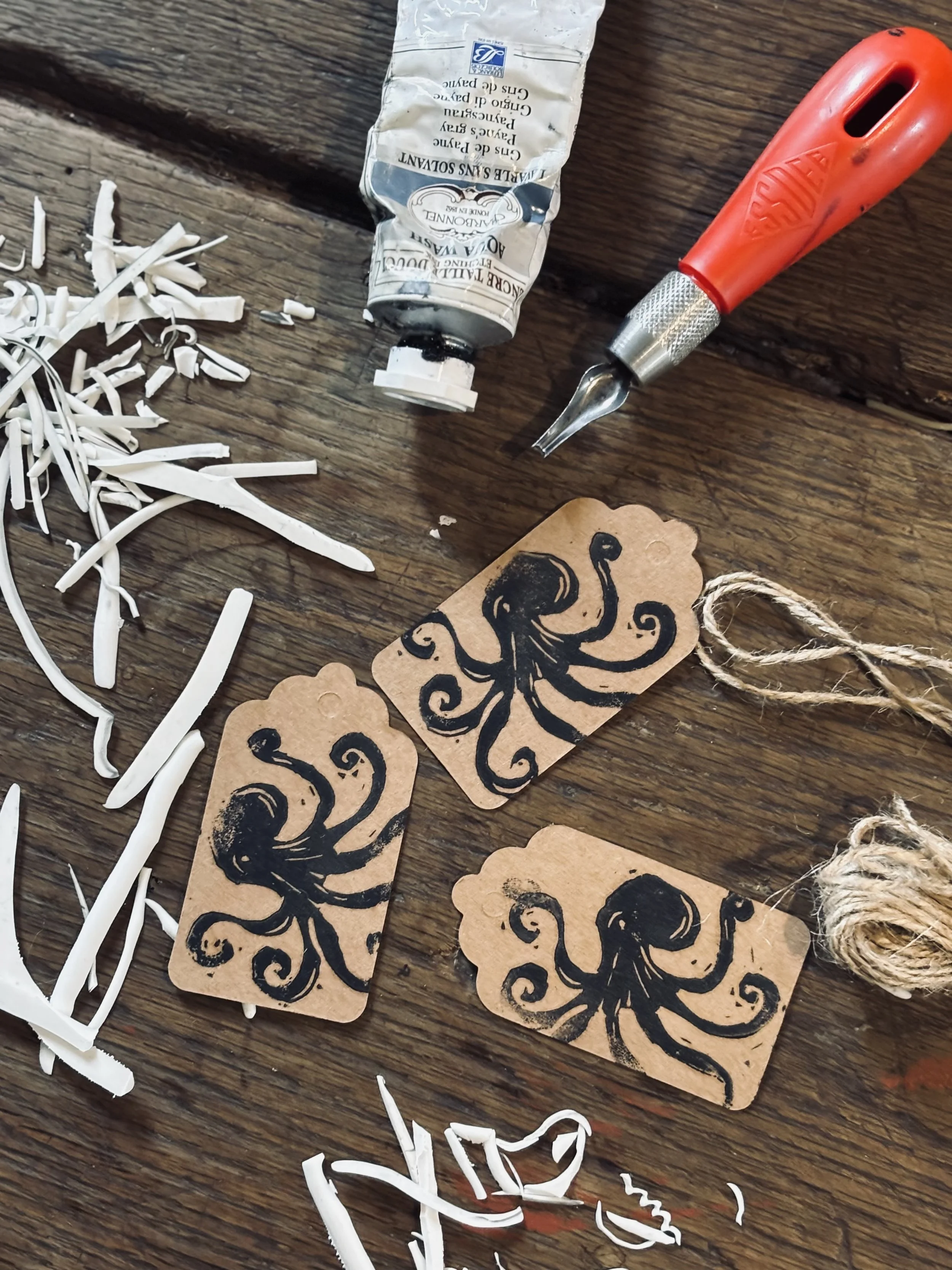 Trois étiquettes en carton avec un dessin d'une pieuvre noire, un tube de colle, un cutter, des morceaux de plastique blanc, une pelote de ficelle.