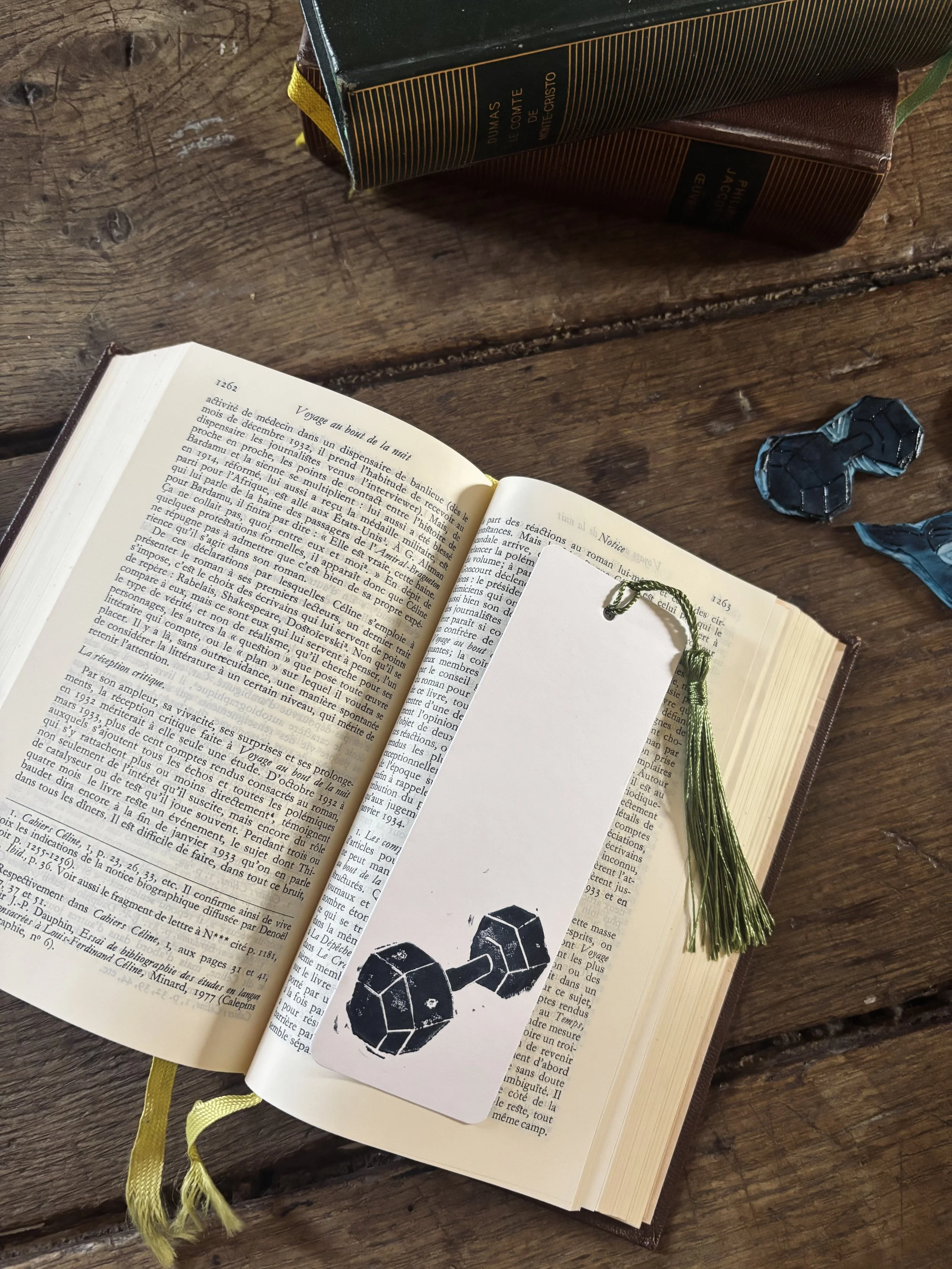 Un livre ouvert avec un signet vert, un marque-page représentant une chaîne de poids, sur une table en bois, avec d'autres livres empilés et un petit objet noir et bleu