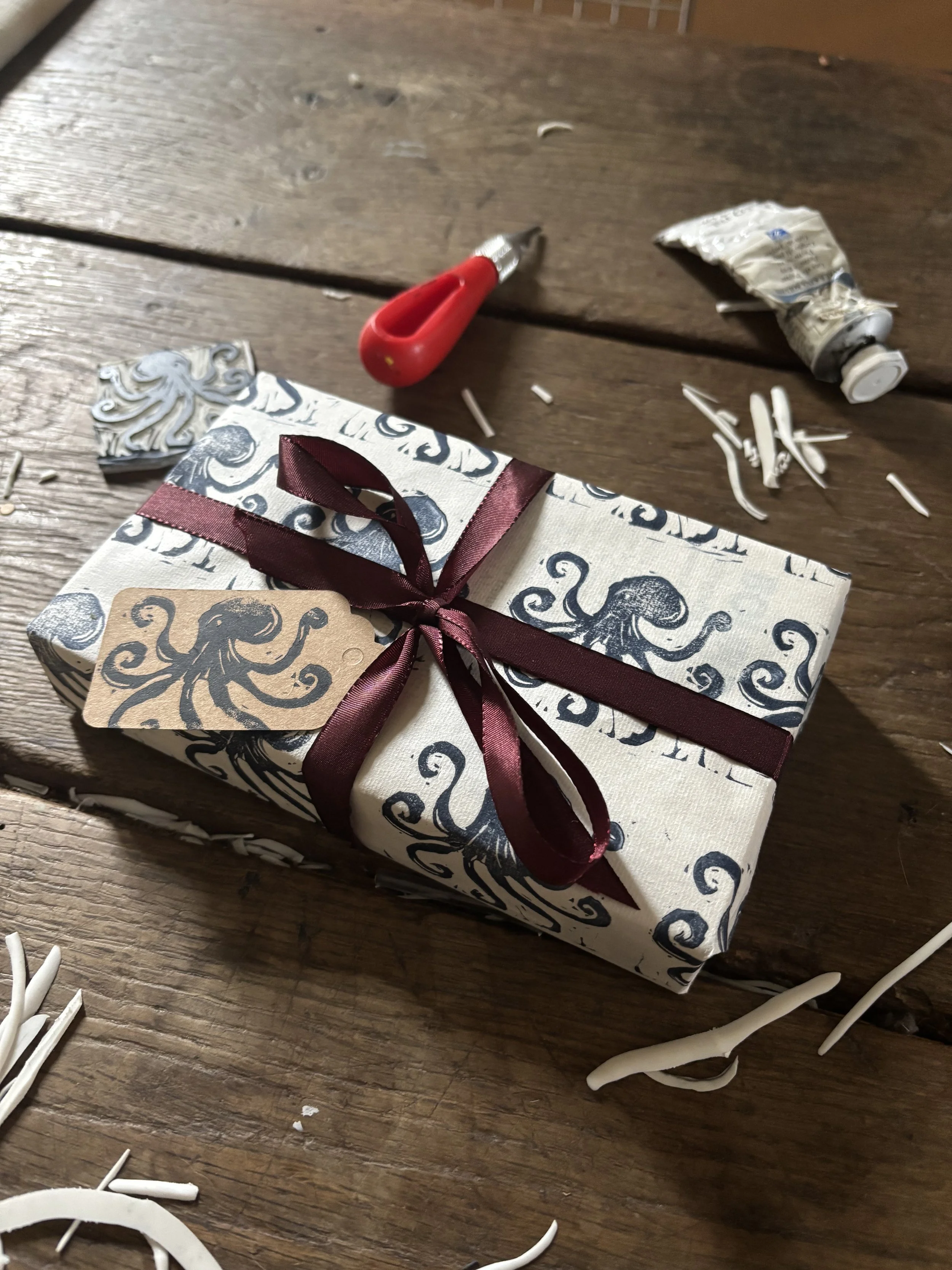Un cadeau emballé avec du papier décoré d'encre noire avec un motif de pieuvres, attaché avec un ruban marronâtre en satin, posé sur une table en bois avec des morceaux de papier d'emballage et un cutter à proximité.