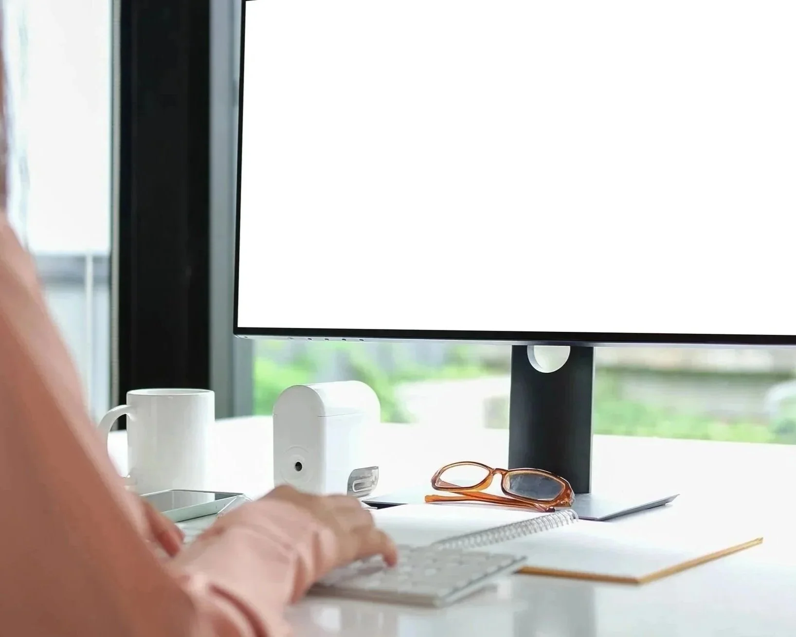 Office workspace with computer monitor, keyboard, notebook, glasses, and a cup on a white desk.