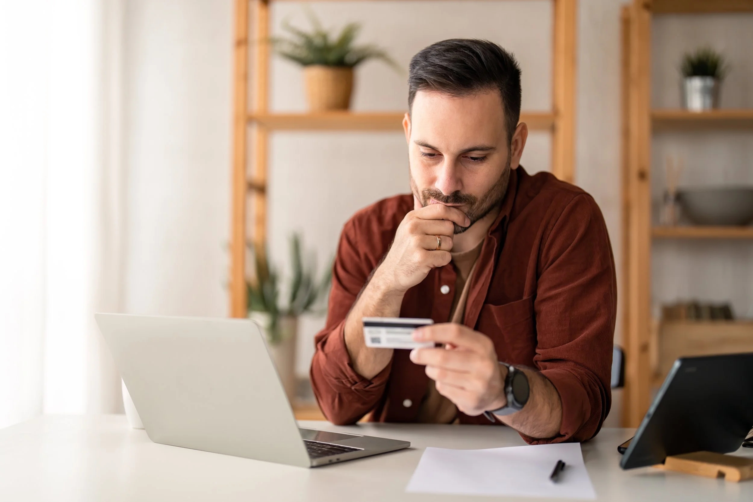 Man looking at a credit card with a laptop, paper, and tablet on a white desk in a home office.
