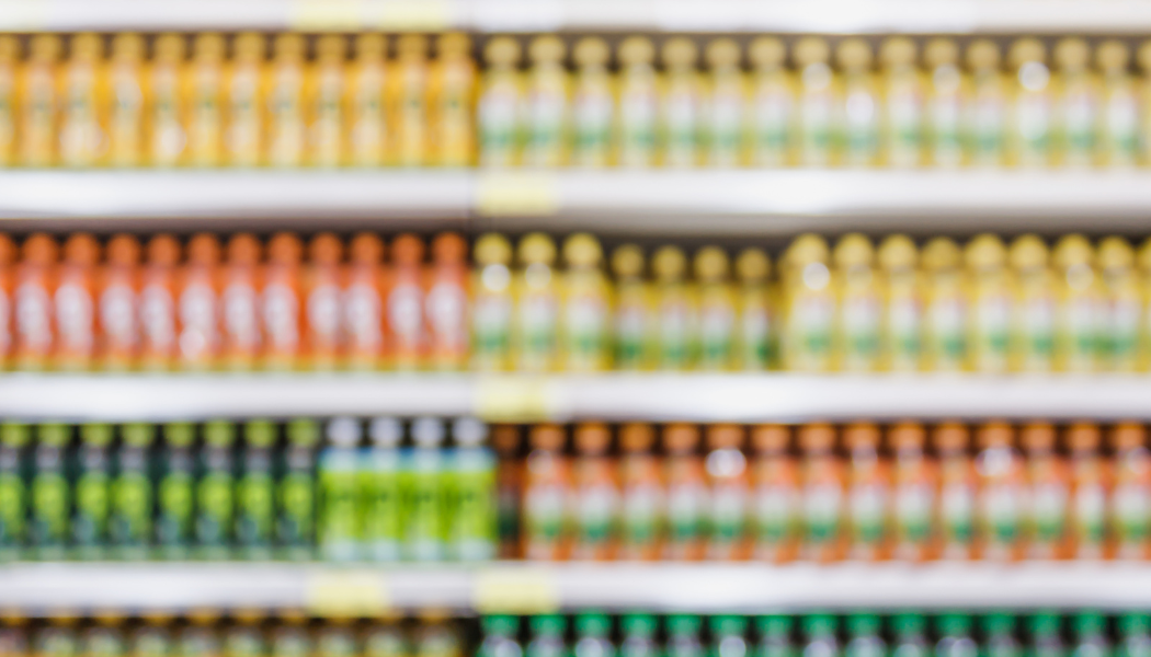 Blurry image of grocery store shelves stocked with various bottles in multiple colors including yellow, orange, and green.