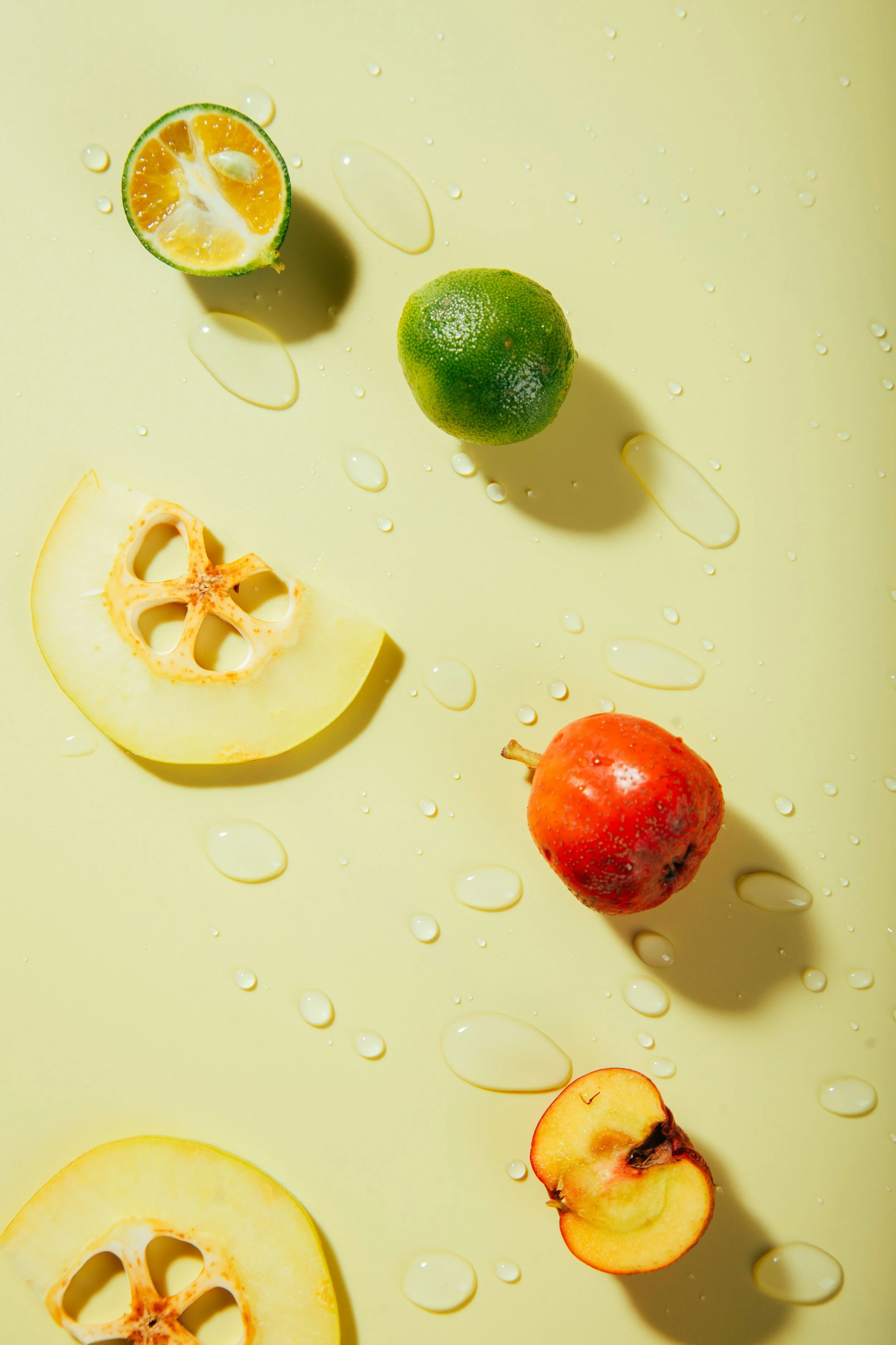 Various fruits including a lime, a halved lemon, a pomegranate, and a halved apple on a yellow background with water droplets.