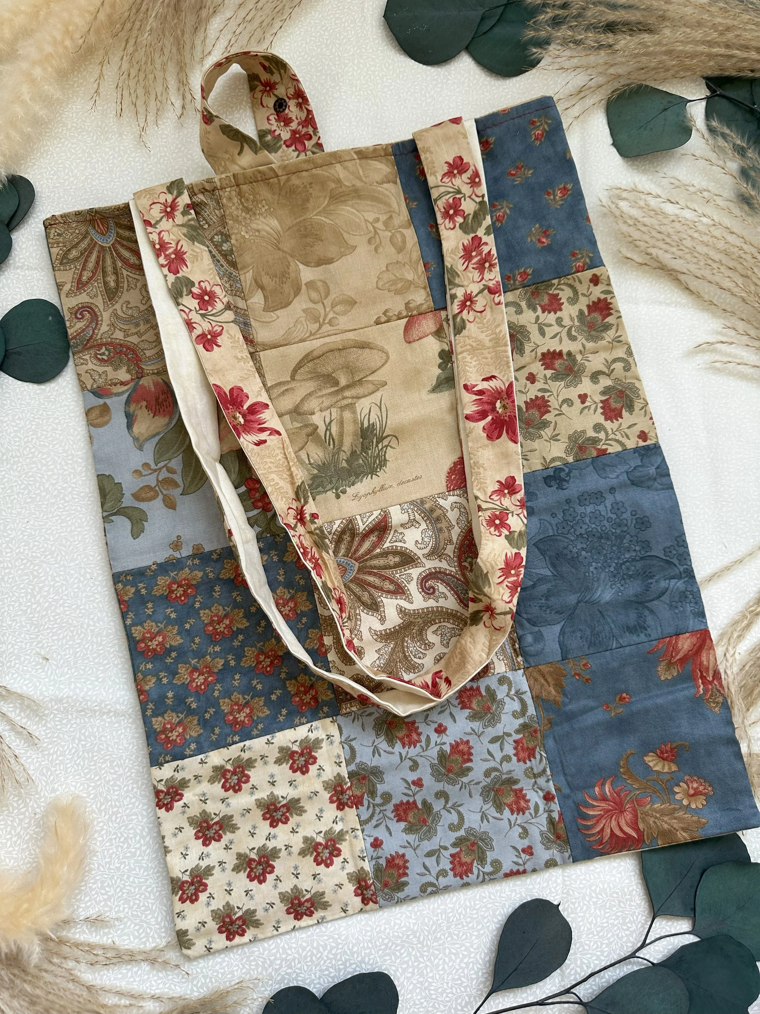 A patchwork tote bag with floral and mushroom designs, placed on a white surface with dried grasses and eucalyptus leaves around it.
