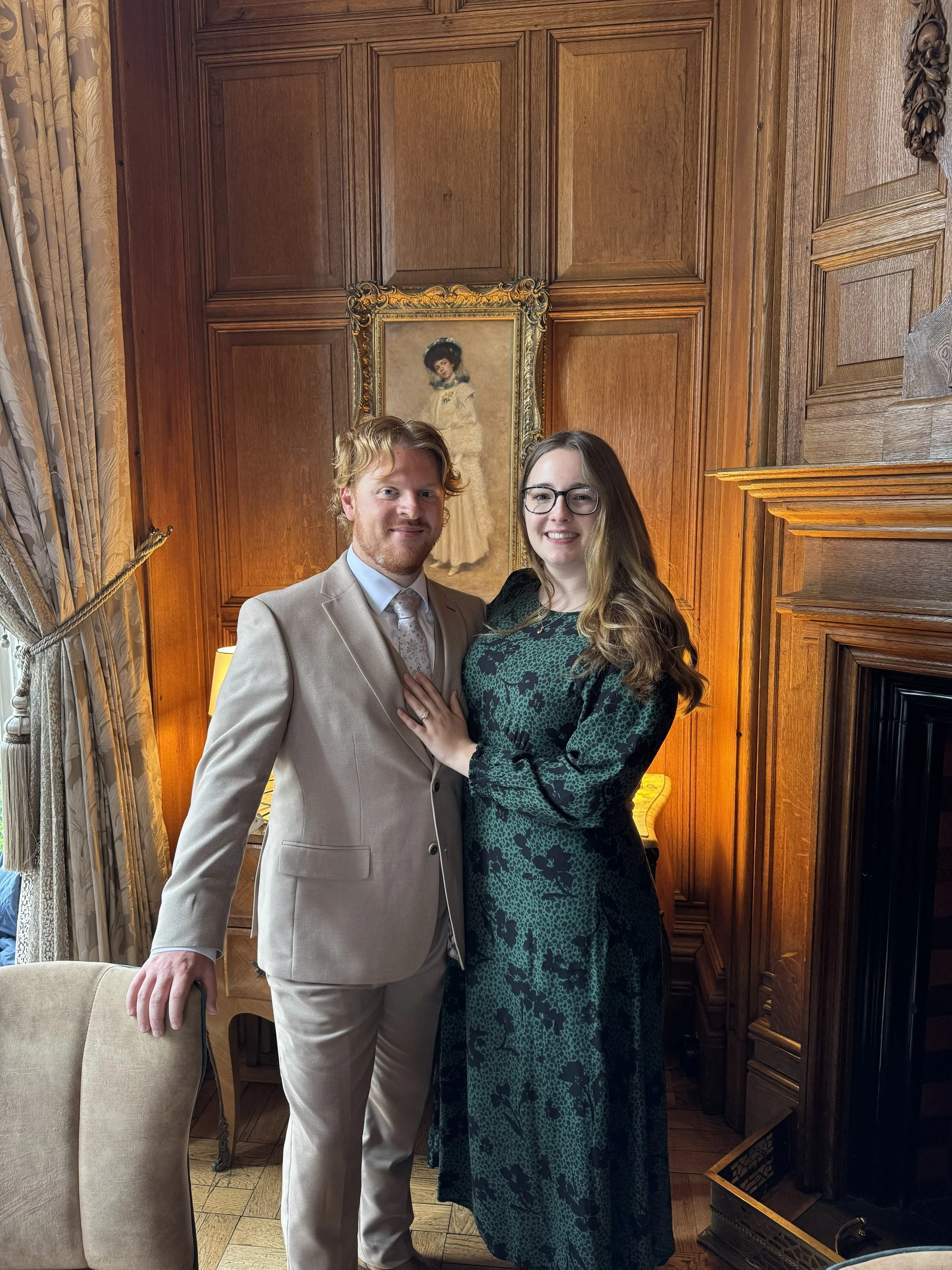 A man and woman standing together in a wood-paneled room, smiling for the camera. The man is wearing a light beige suit with a white shirt and patterned tie, and the woman is dressed in a long, dark green and black patterned dress with glasses. There is a framed portrait of a woman in a vintage dress hanging behind them, an ornate curtain on the left, and a wooden fireplace on the right.