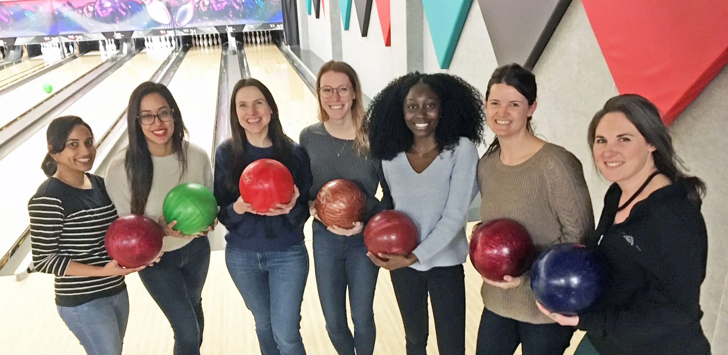 A group of seven women at a bowling alley, each holding a colorful bowling ball, and smiling