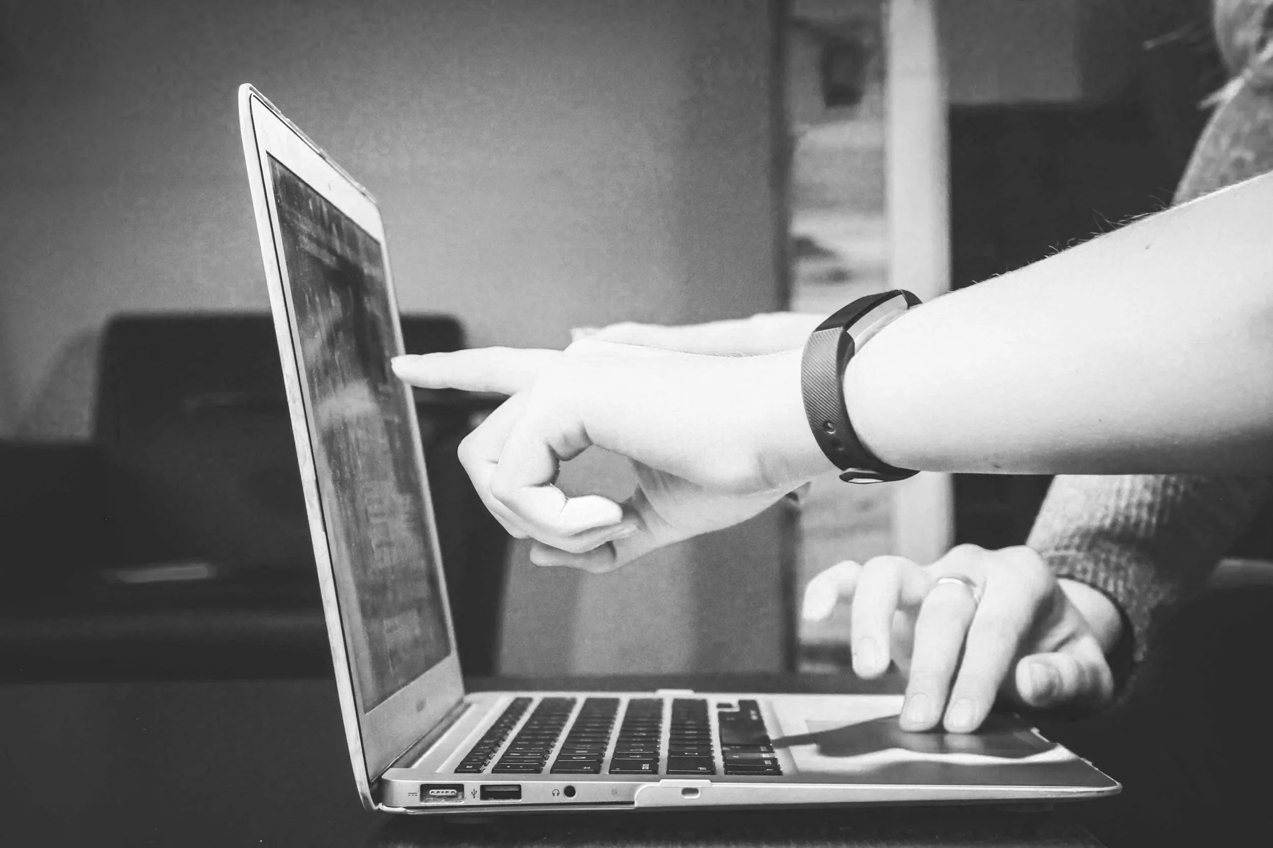 A person interacts with a laptop while another person points at the screen, in grayscale.