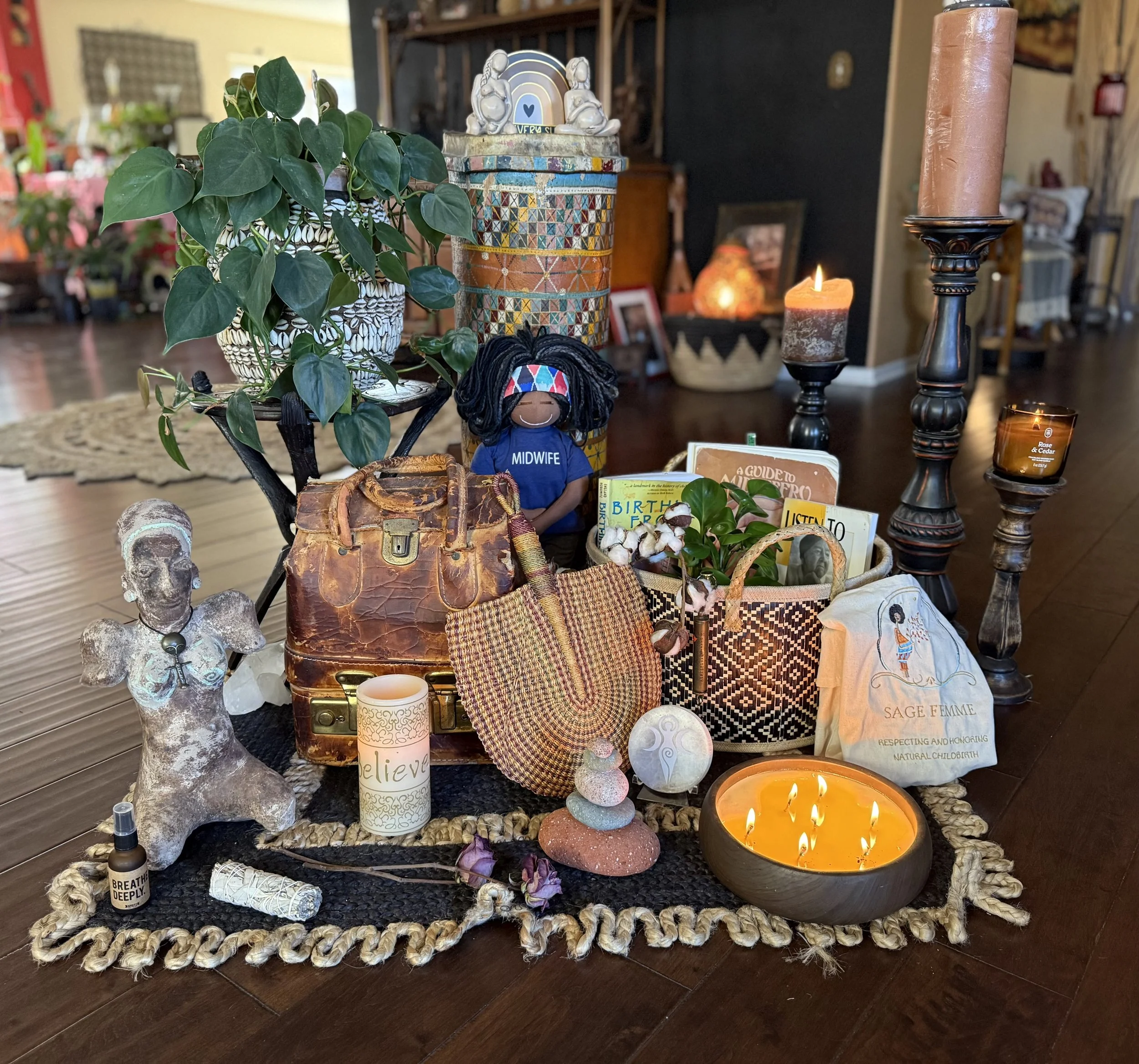 Decorative arrangement on a table featuring a potted plant with large green leaves, small figurines, candles, books, and various ornaments, set on a woven mat with a dark hardwood floor underneath.