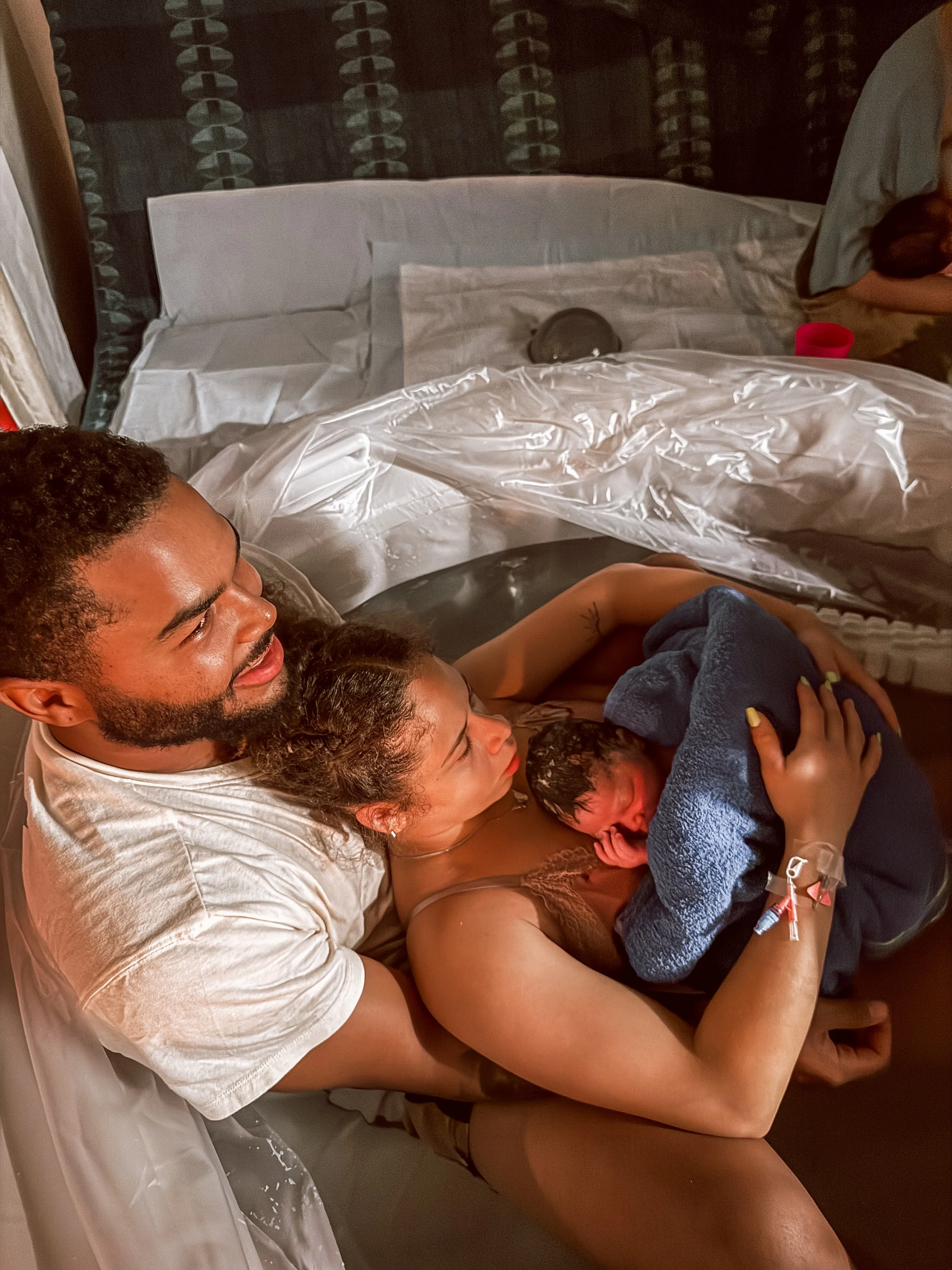 A man, woman, and baby in a hospital delivery room, after childbirth. The man is smiling, the woman is lying down with the baby on her chest, and a small blanket is covering the baby's body.