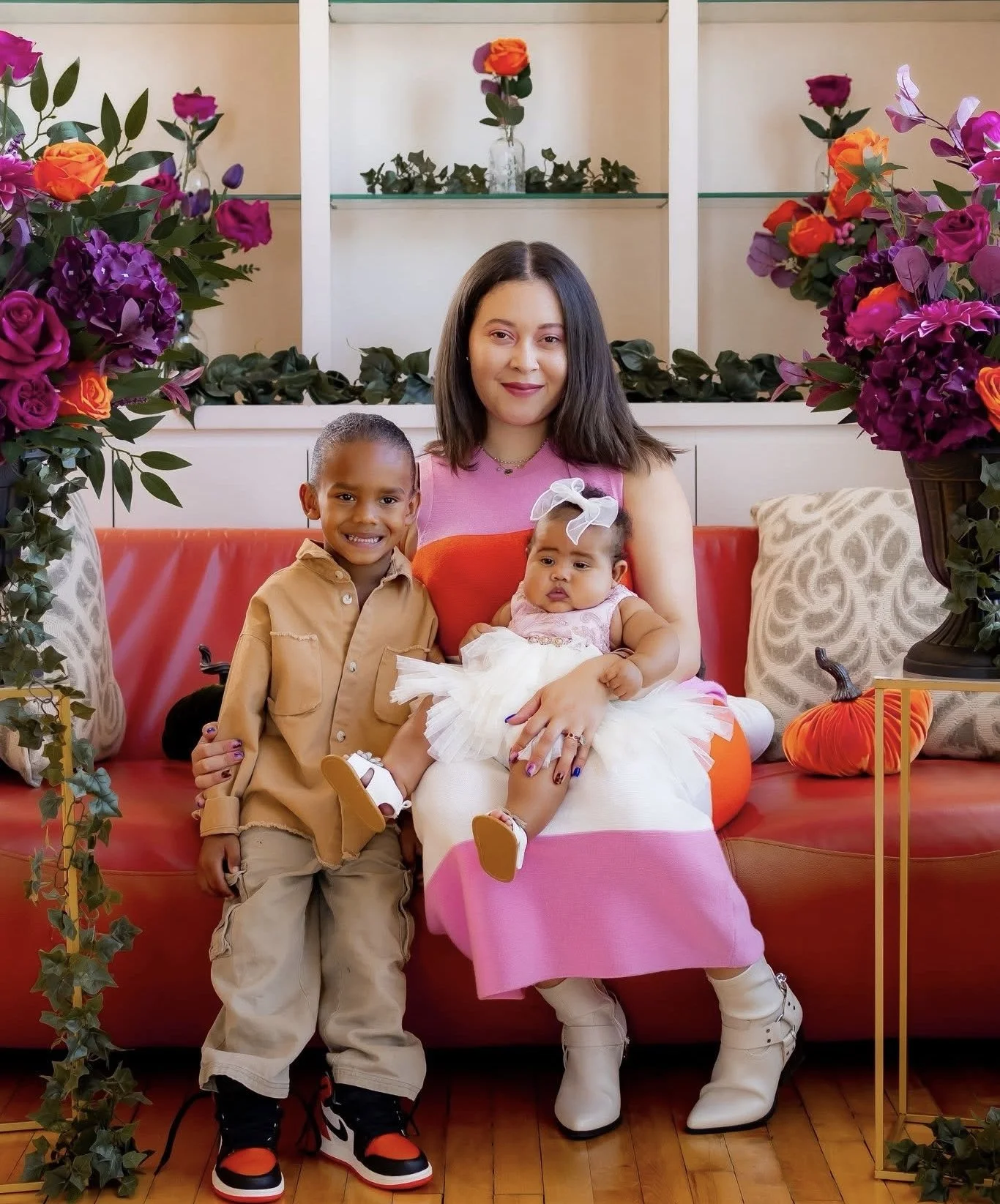 A woman with shoulder-length brown hair wearing a pink and orange dress, sitting on a red sofa with two children. The boy is standing next to her, smiling, wearing a beige shirt and beige pants. The girl is sitting on her lap, wearing a white dress with pink accents, white boots, and a white bow in her hair. The background has white shelves with glass vases holding flowers, and there are large floral arrangements with purple, pink, and orange flowers on either side of the sofa. The setting appears to be decorated for a festive occasion.
