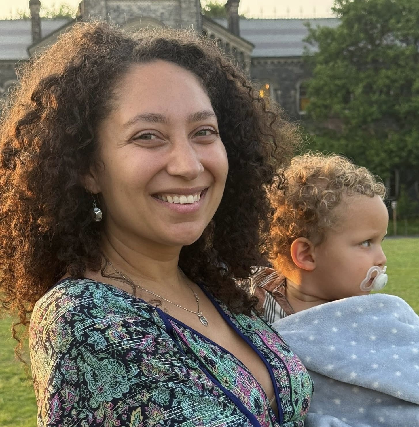 A woman with curly hair smiling and holding a young child with curly hair and a pacifier outdoors in a park.