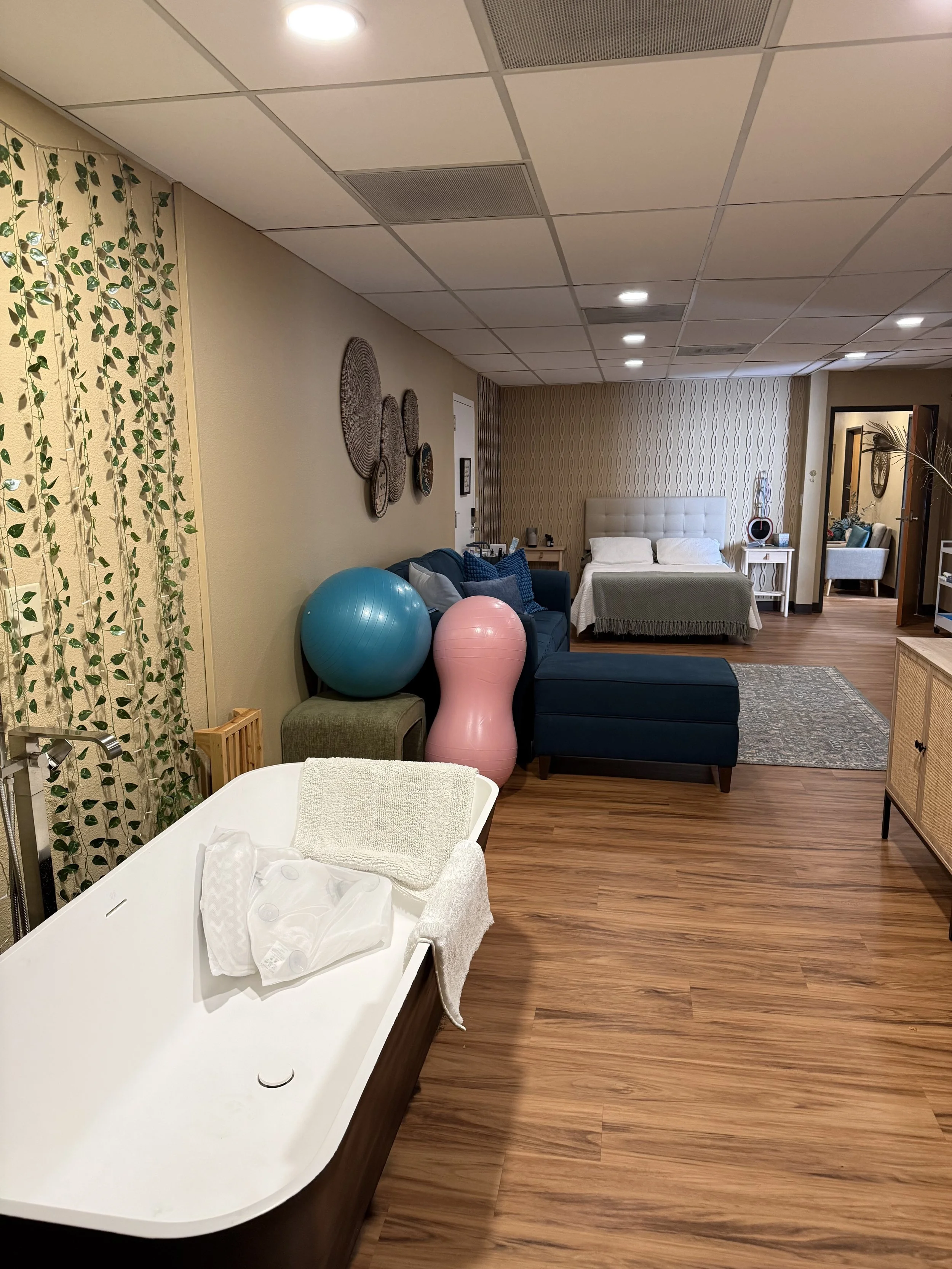 A hospital room with a baby bassinet, a changing table, and a bed with pillows, with exercise balls on a couch and decorative wall art.