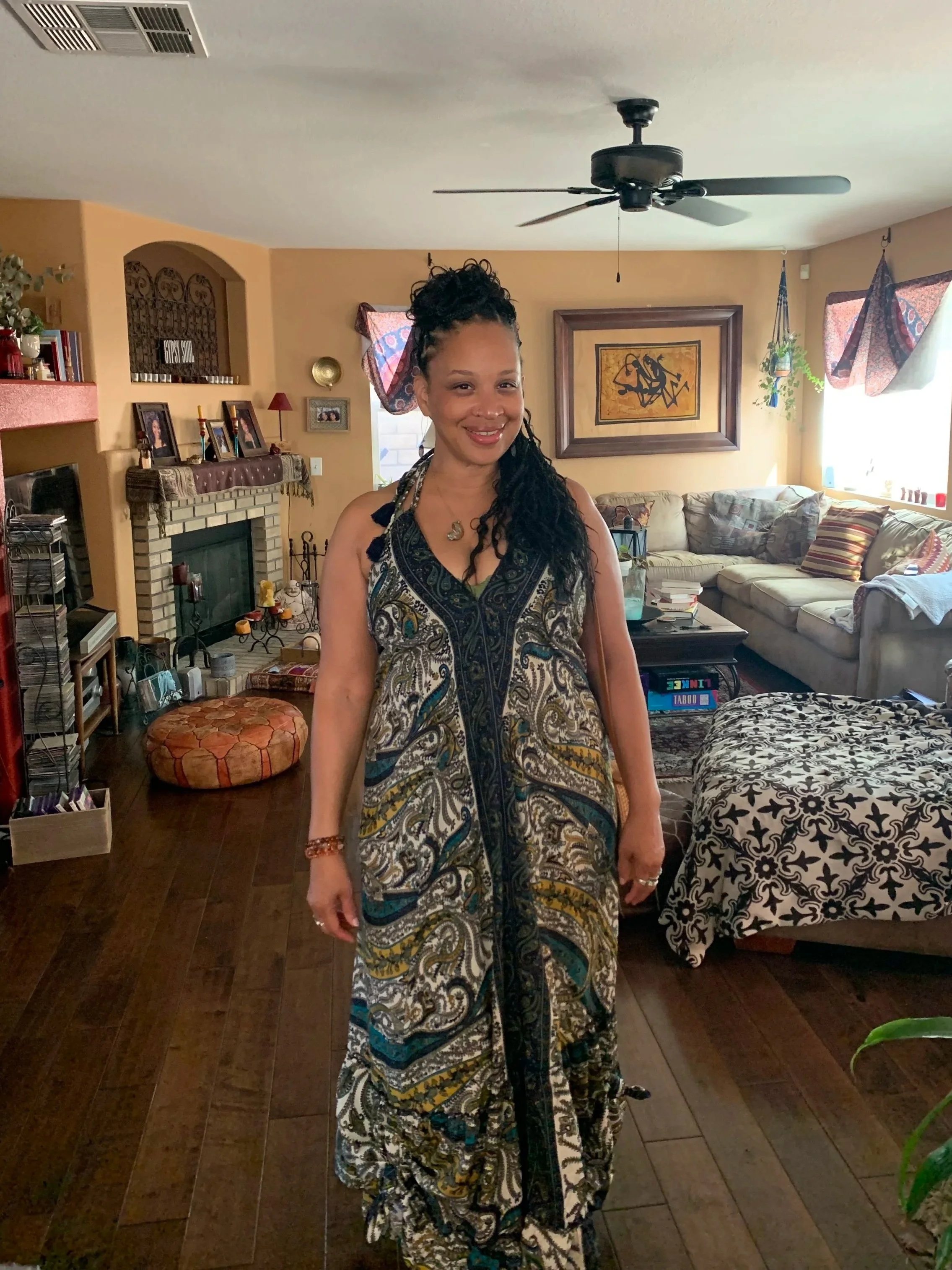 A woman with curly black hair styled in a high bun, wearing a bohemian patterned dress, and standing in a living room with beige walls, a brick fireplace, and a beige sectional sofa.