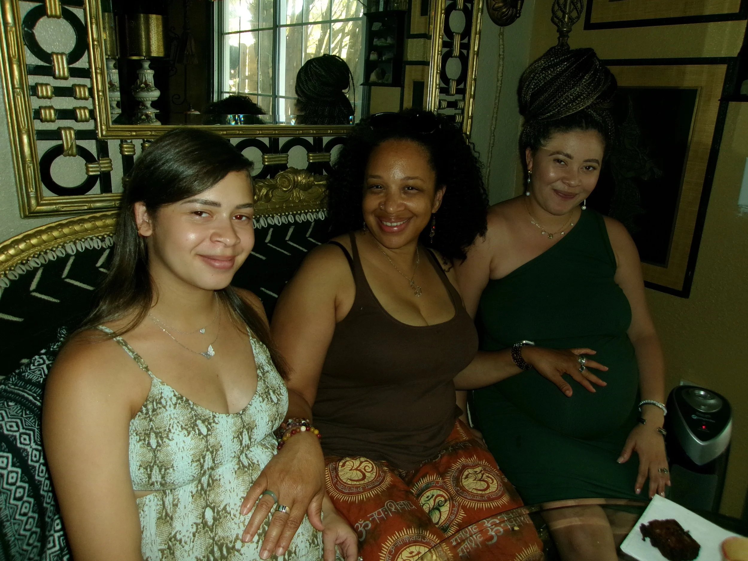 Three women sitting together in a room with ornate black and gold furniture, a large mirror behind them, and a window with natural light. They are smiling and dressed in casual, colorful clothing.
