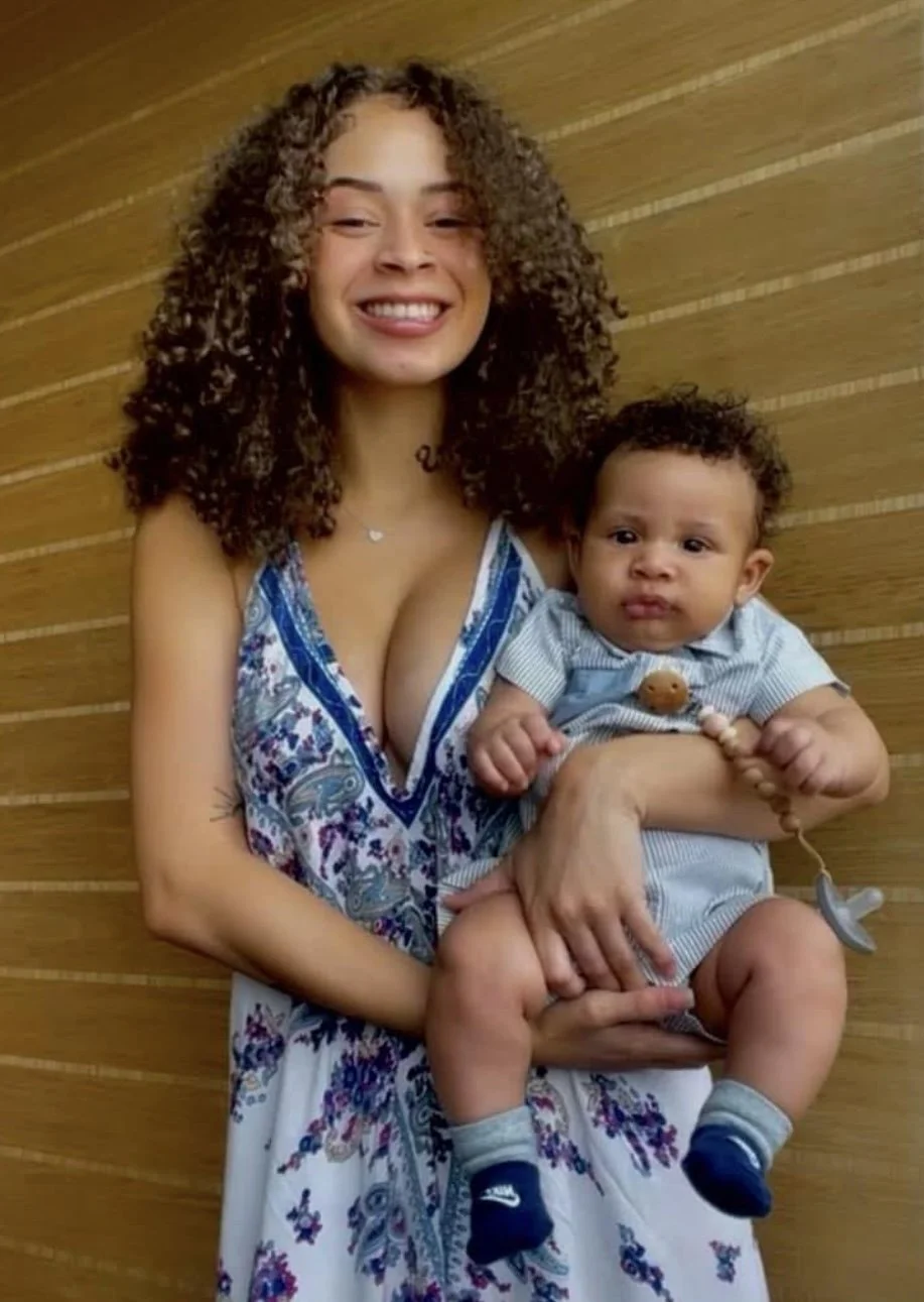 A woman with curly hair holding a young child with short curly hair against a wooden wall background, both smiling.
