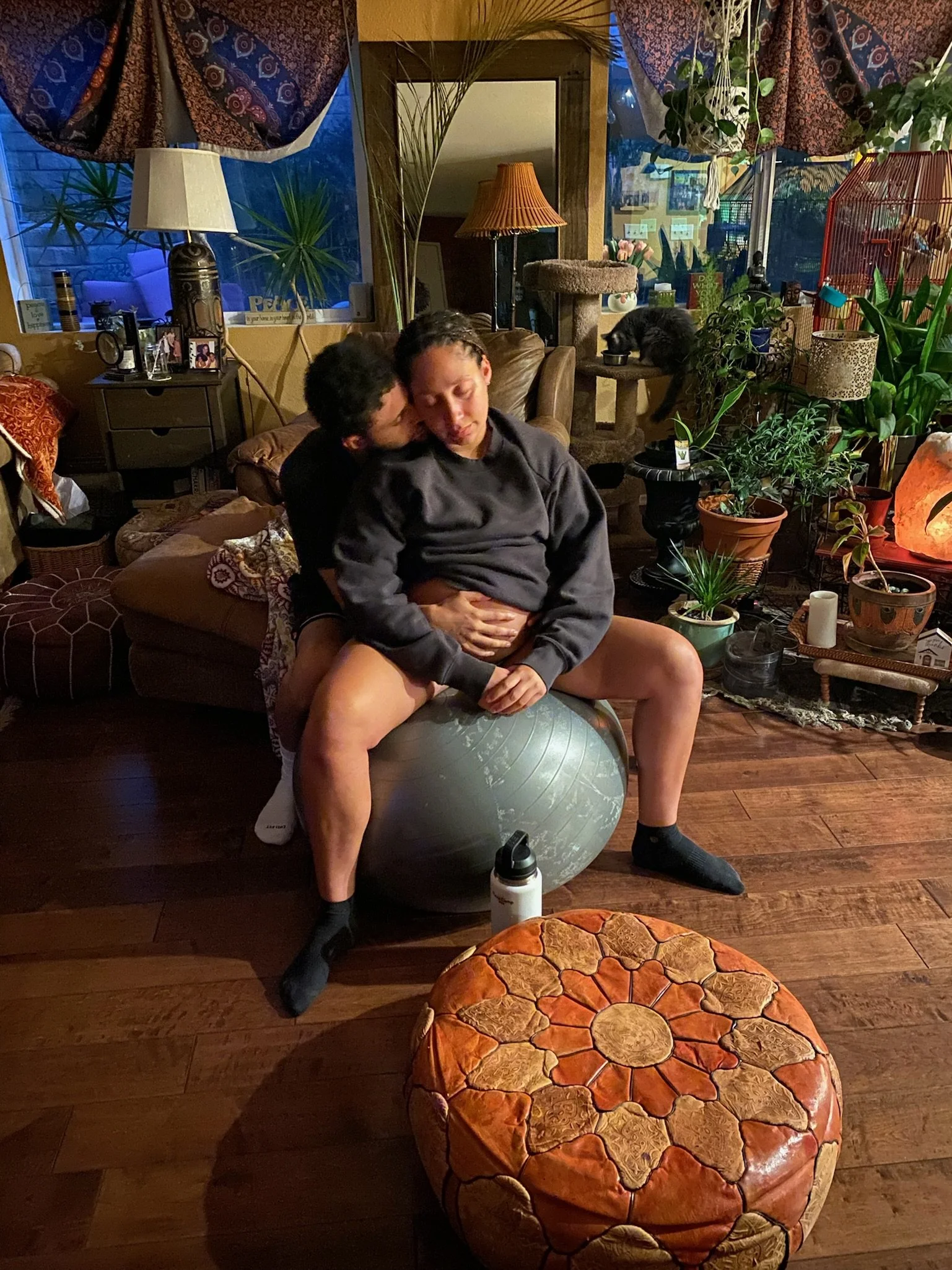 Two people are sitting on a yoga ball on a living room floor, with one person in front and the other behind, in a cozy and decorated room filled with plants and miscellaneous items.
