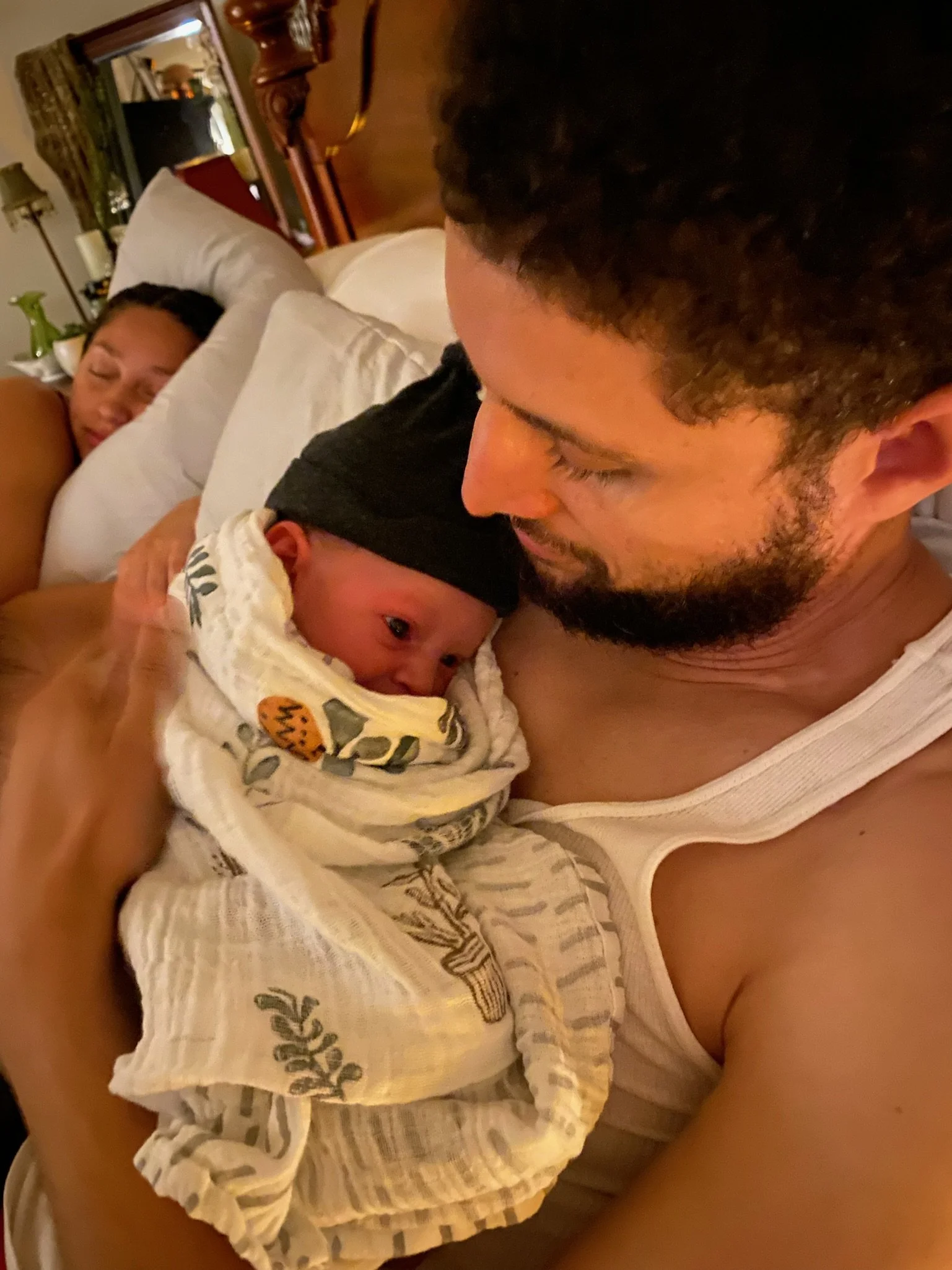 A man holding a newborn baby close to his face, with a woman sleeping in the background on a bed in a cozy room.