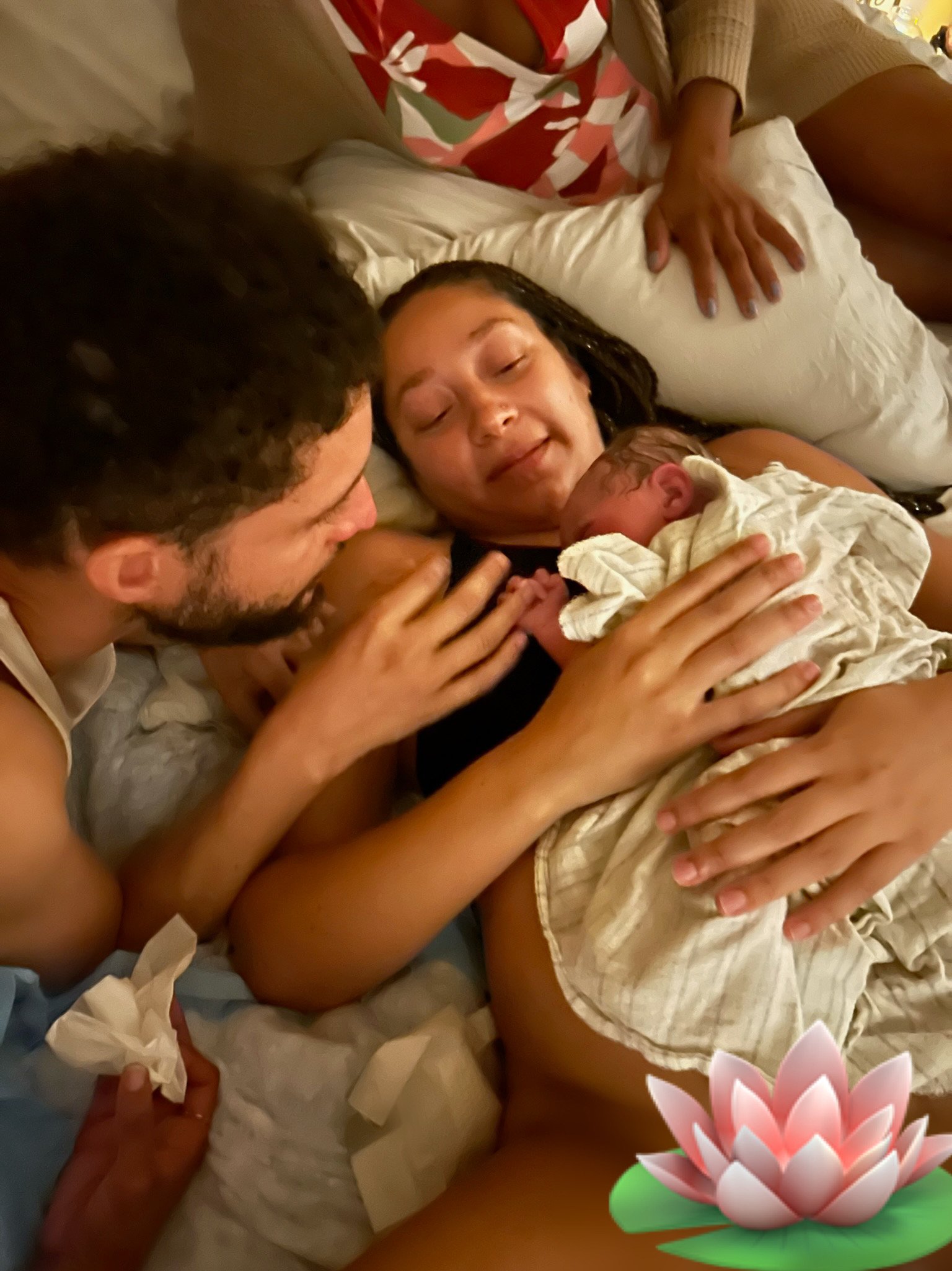 A woman lying in bed holding a newborn baby, with a man and another child's hands visible. The image has a lotus flower and a pink flower overlay.