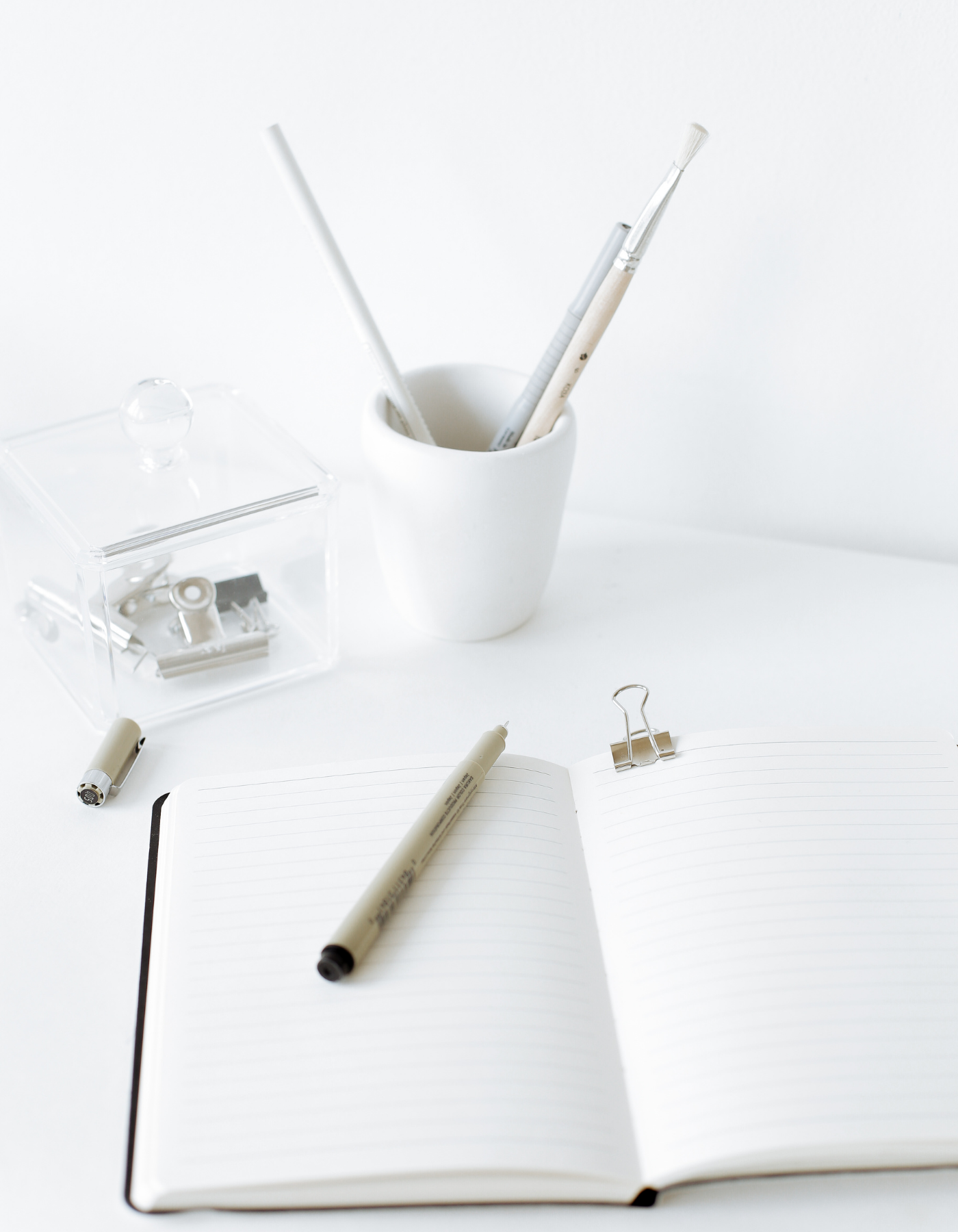 A white open notebook with a black cover, a beige marker on pages, and a gold paperclip, surrounded by white pens, a clear box with clips, and a white cup with three brushes, set on a white background.
