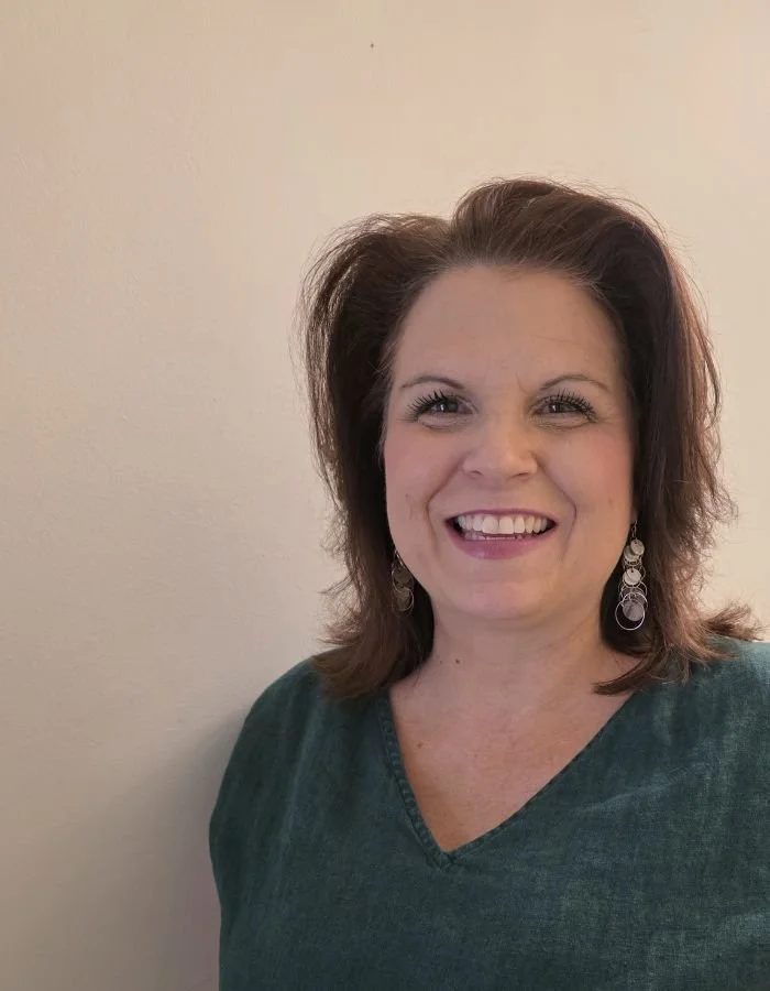 A woman smiling with shoulder-length brown hair, wearing earrings and a green top, standing against a plain light-colored wall.