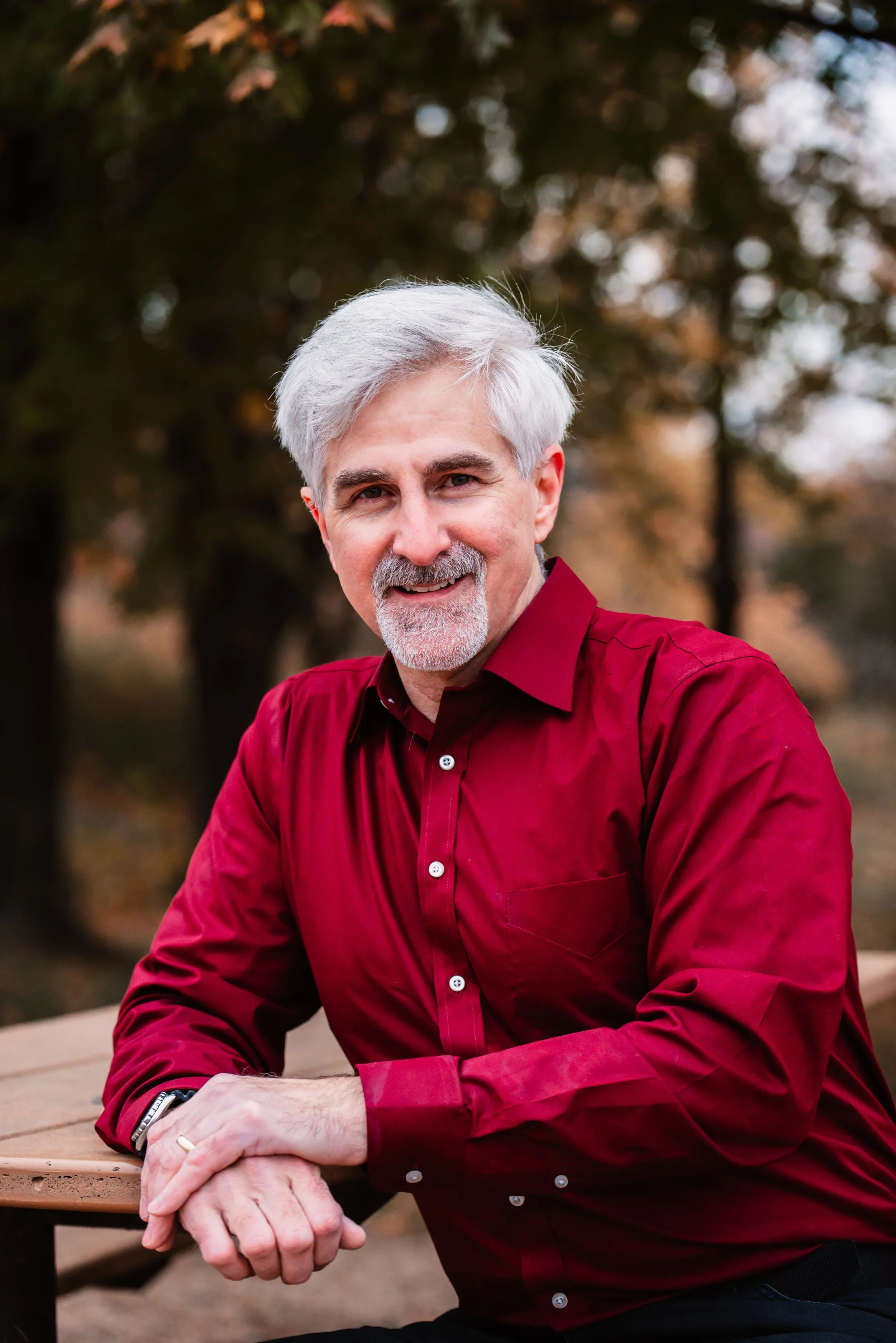 A middle-aged man with gray hair, gray beard, and a friendly smile, wearing a red button-up shirt, sitting outdoors at a wooden table with a background of trees and autumn foliage.