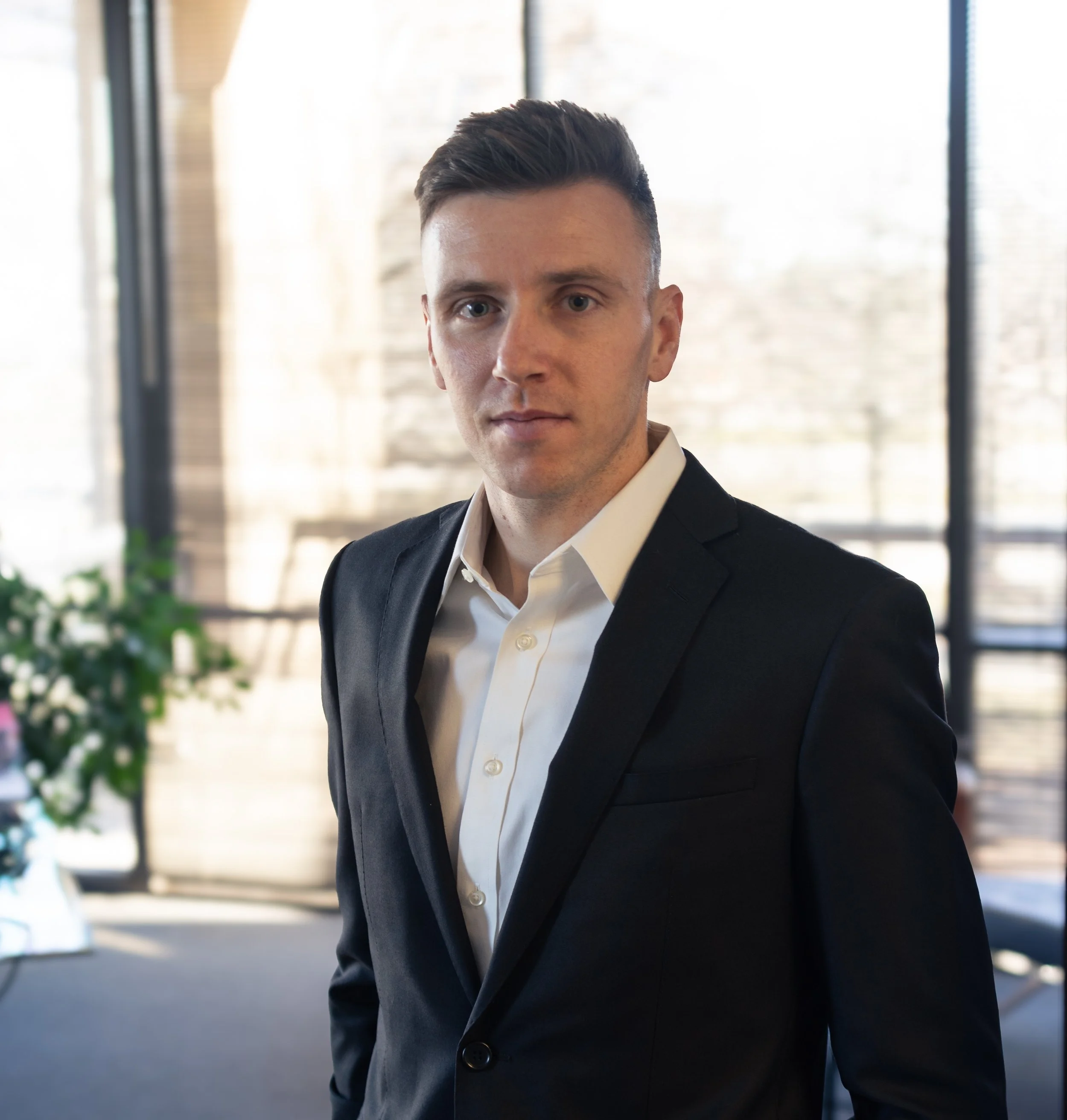 A young man in a black suit and white shirt standing in an office with large windows and some greenery.