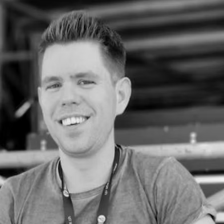 Ein junger Mann mit kurzen, dunklen Haaren, lächelnd, im Freien, schwarz-weiß, trägt ein T-Shirt und eine Lanyard.