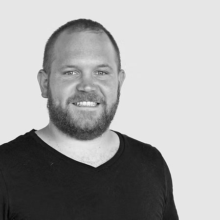 Schwarz-weiß-Foto eines Mannes mit kurzem Haar, Bart und einem schwarzen T-Shirt, der lächelnd in die Kamera schaut.