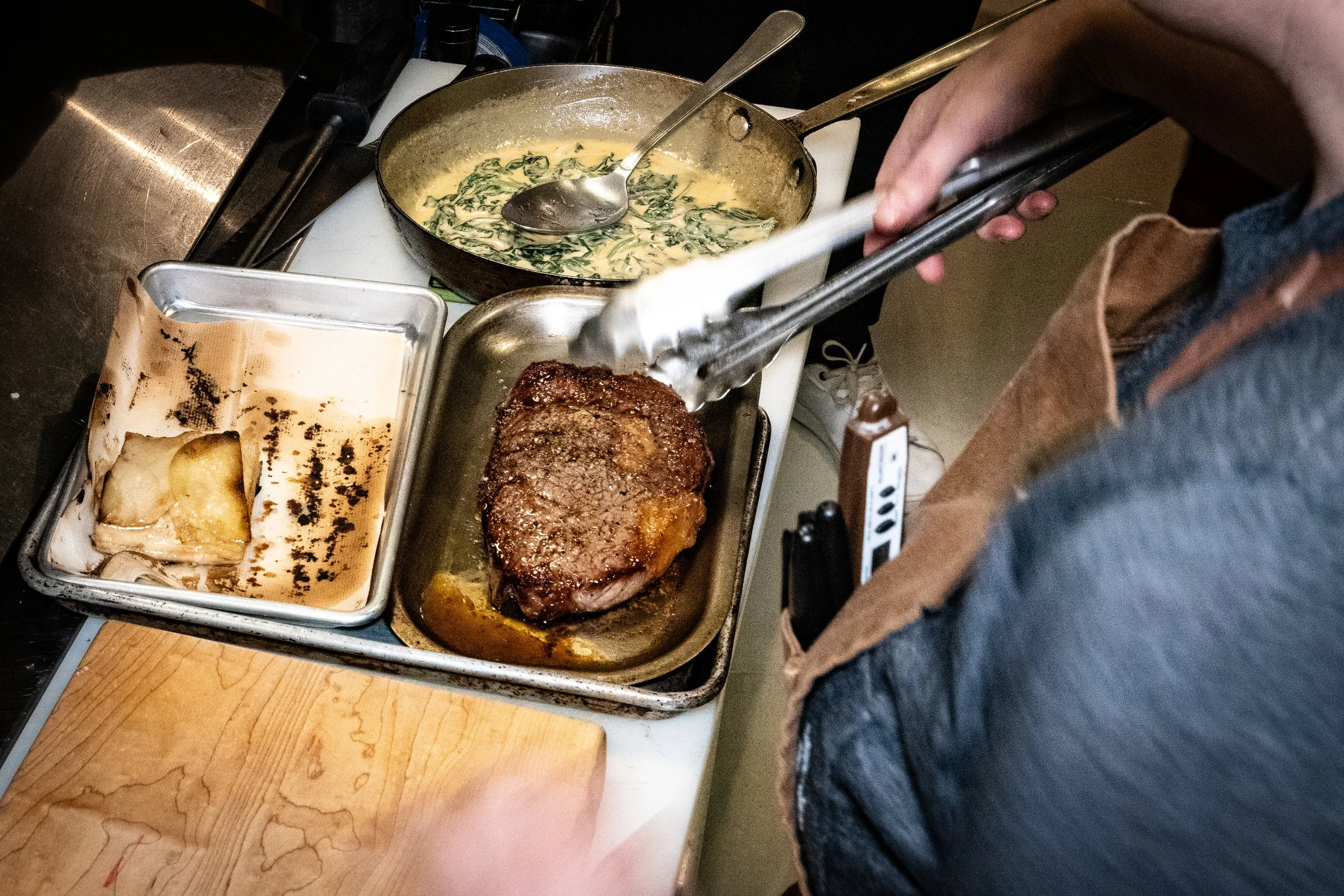Seared steak being transferred with tongs from a roasting pan to a plate, with a baking dish of roasted potatoes and a skillet of creamy spinach in the background.