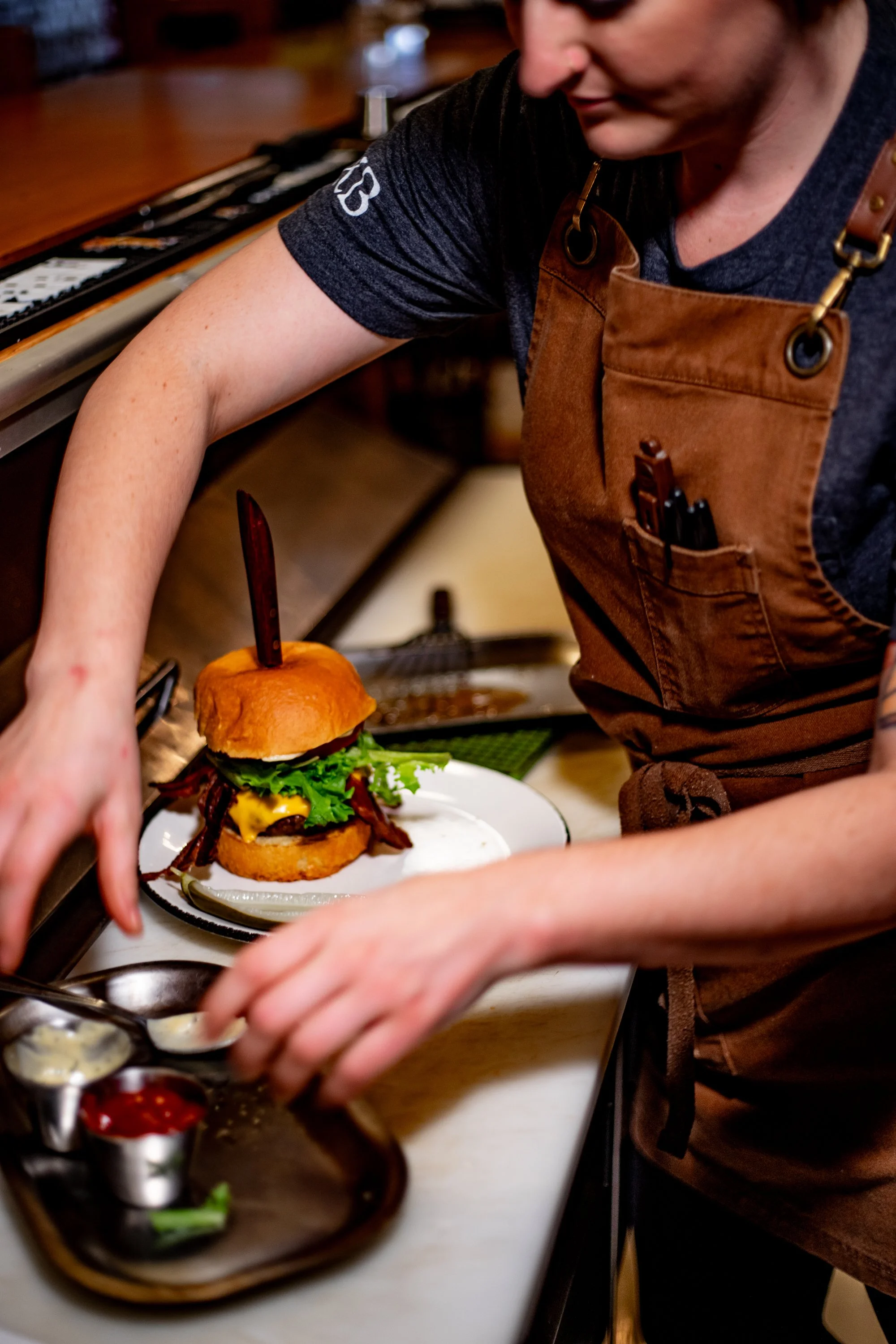 A chef in a brown apron is preparing a gourmet burger with bacon, lettuce, cheese, and tomato on a plate, with various sauces in small containers nearby.