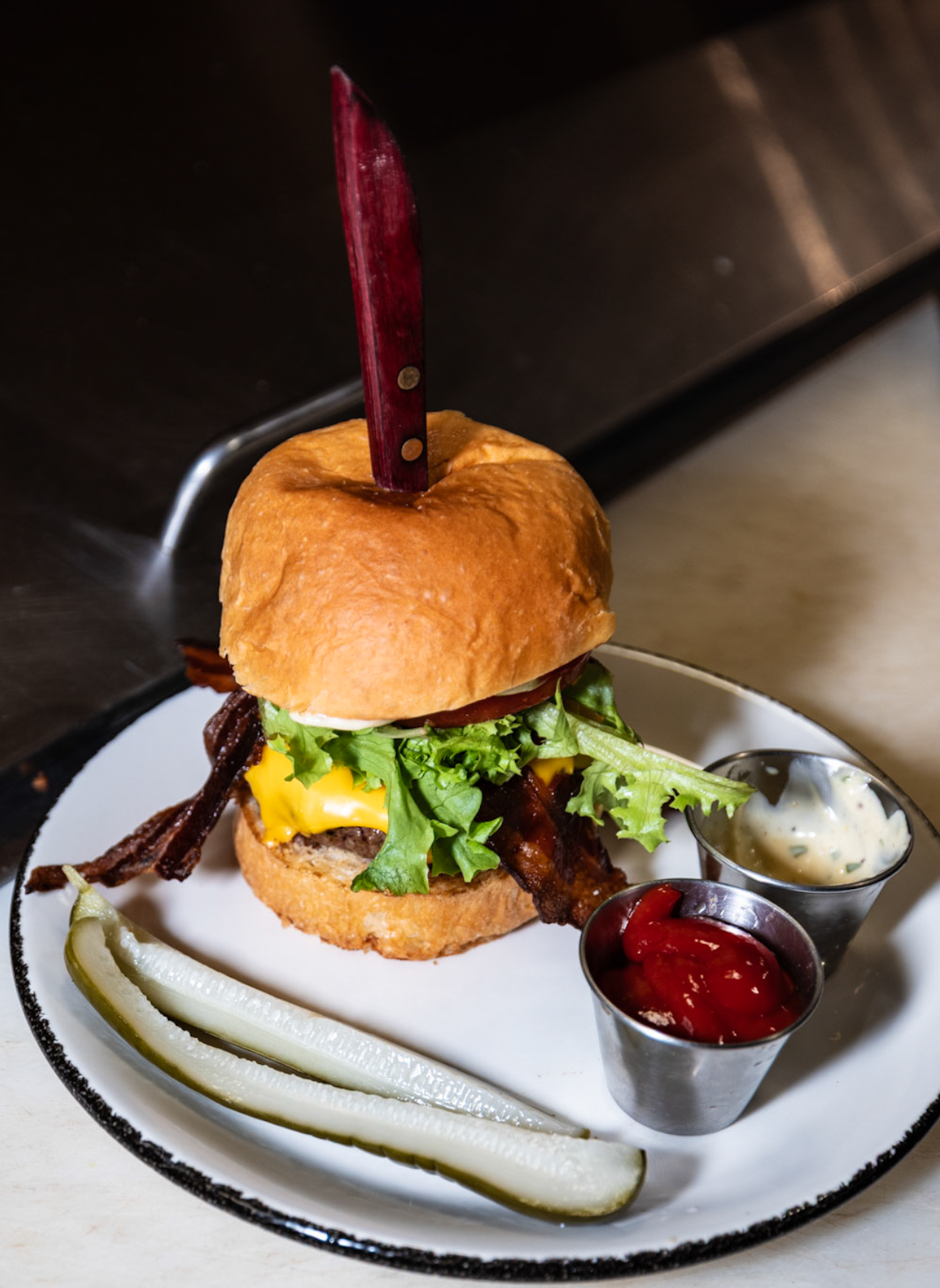 Burger with bacon, lettuce, cheese, tomato, and onion rings, served with ketchup and mayonnaise, with a pickle on the side.