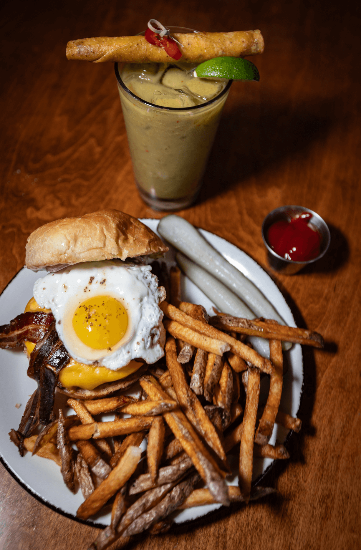 A plate with a burger topped with a fried egg, bacon, and melted cheese, accompanied by French fries and two white sausages. A tall glass of a yellow blended drink with a lime wedge and a rolled wafer cookie is in the background, along with a small container of ketchup.