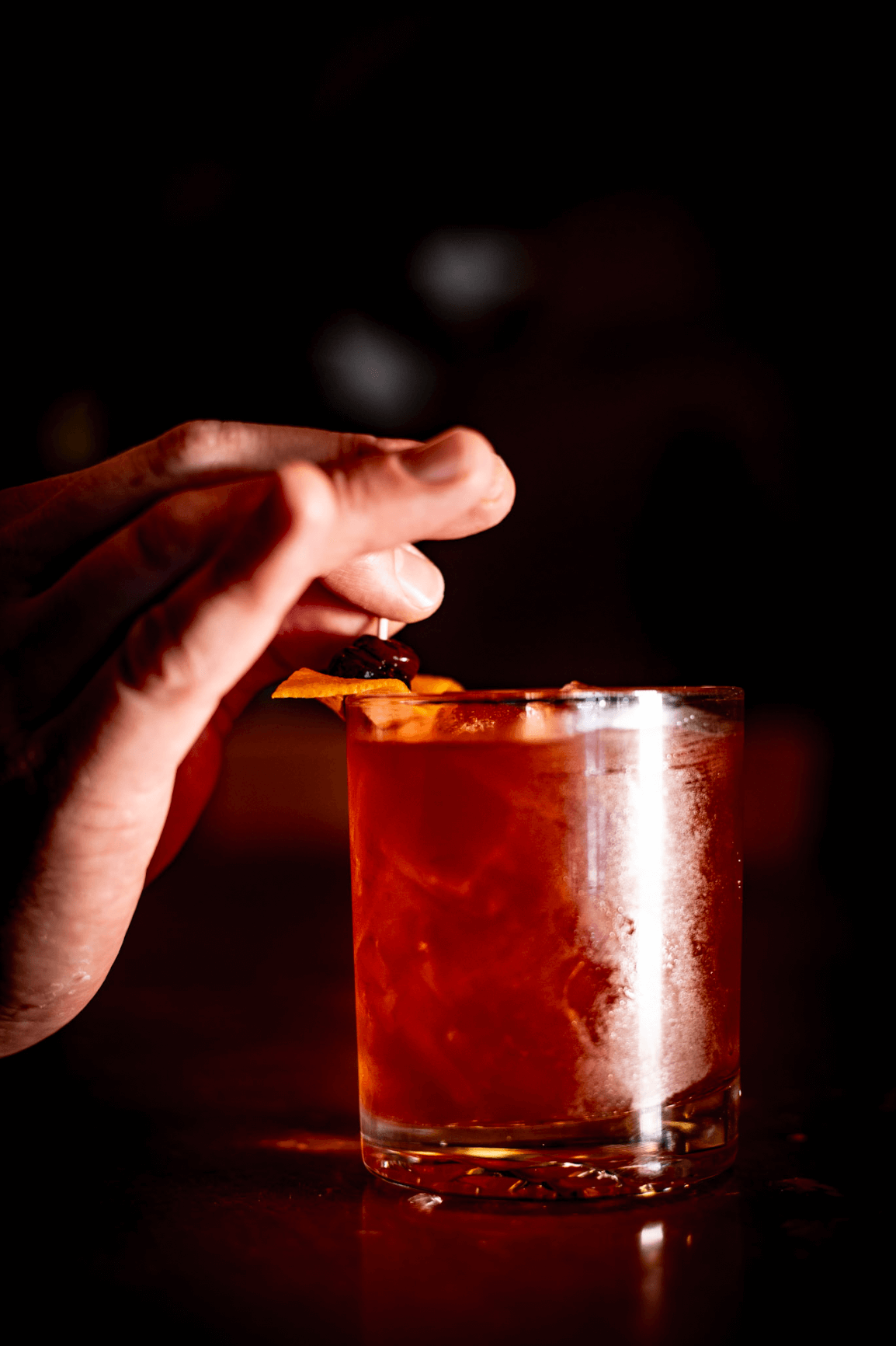 Close-up of a hand garnishing a reddish cocktail with an orange peel and cherry, dark background.