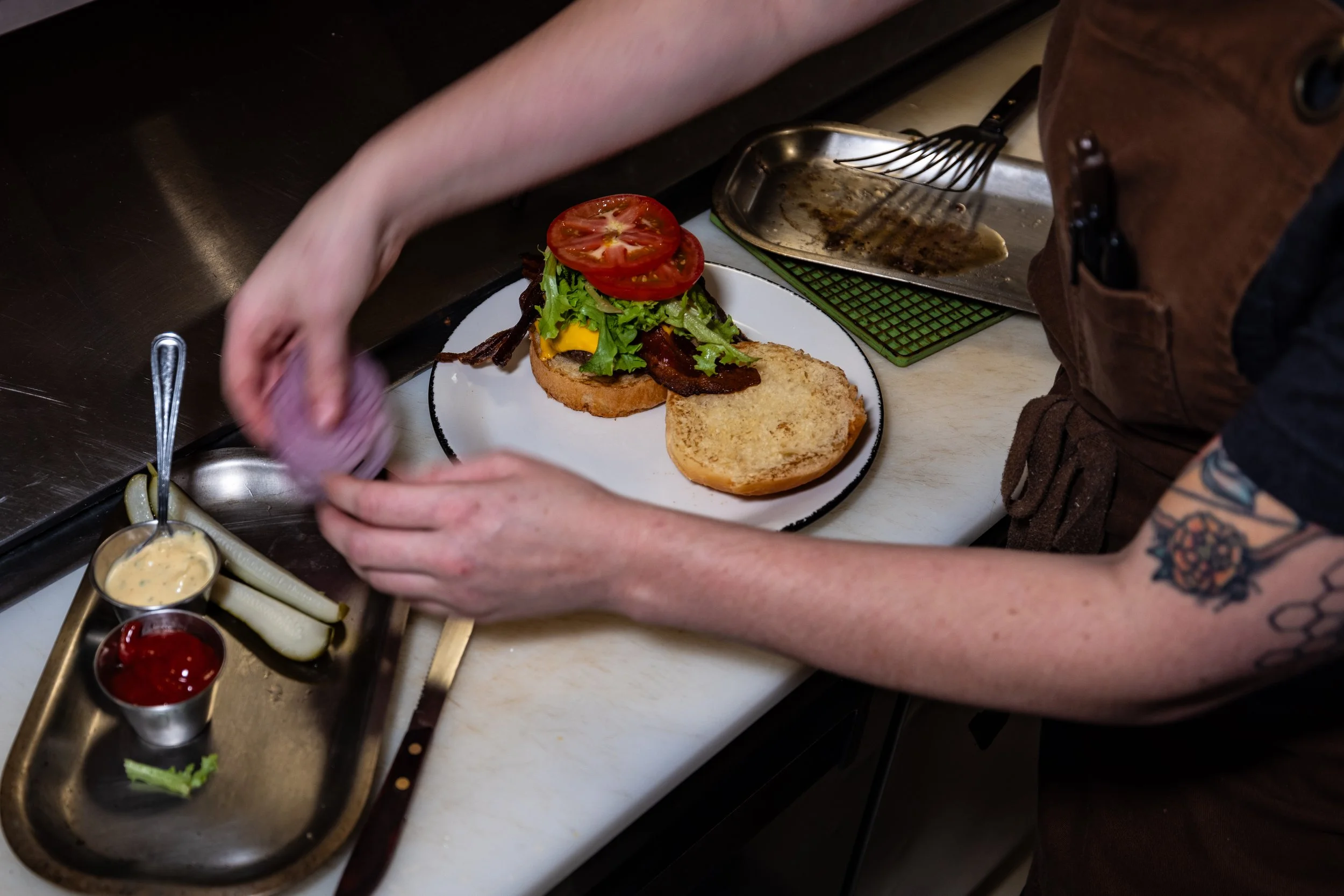 A person preparing a burger with lettuce, tomato, cheese, bacon, on a toasted bun in a kitchen. There are condiments and pickles nearby.