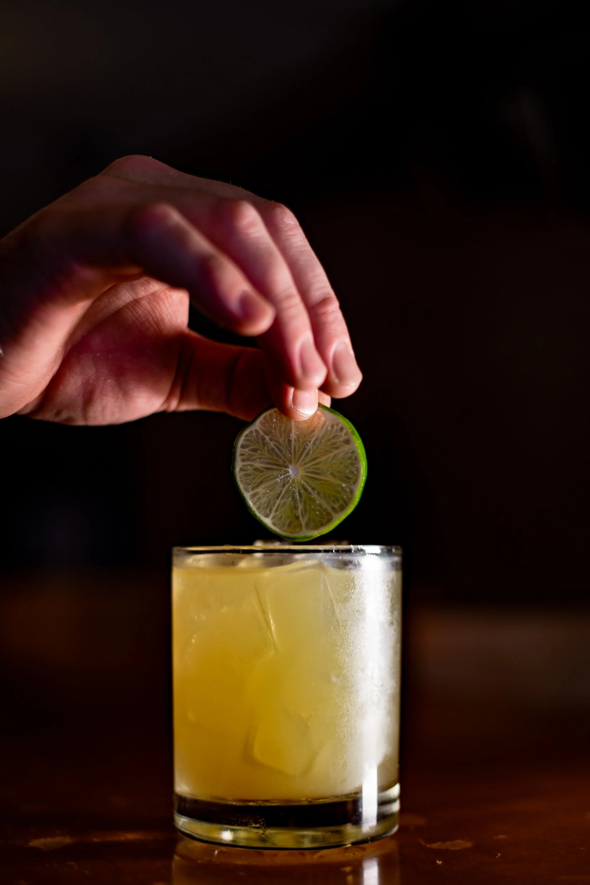 A hand squeezing a lime slice into a glass of yellow cocktail with ice cubes.