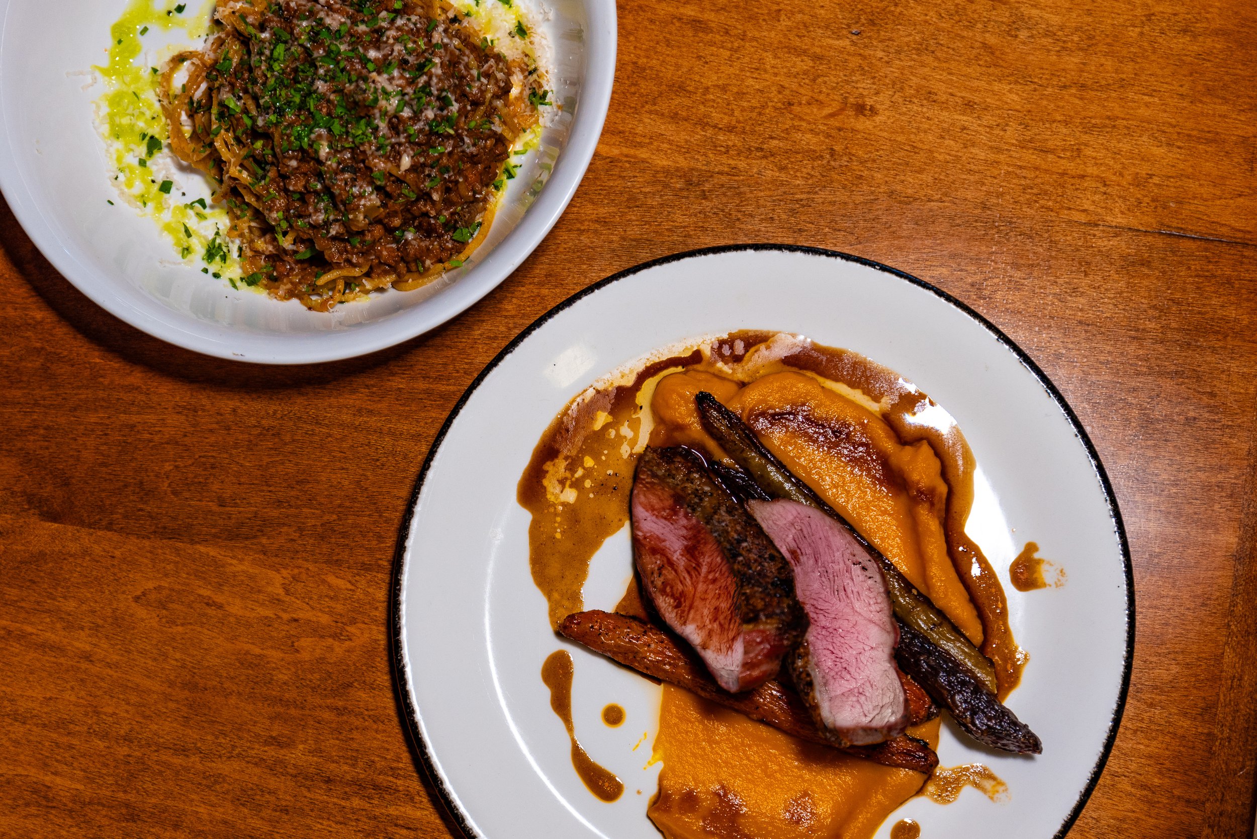 A plate with sliced medium-rare steak, roasted vegetables, and sauce, alongside a bowl of pasta with meat sauce and herbs on a wooden table.