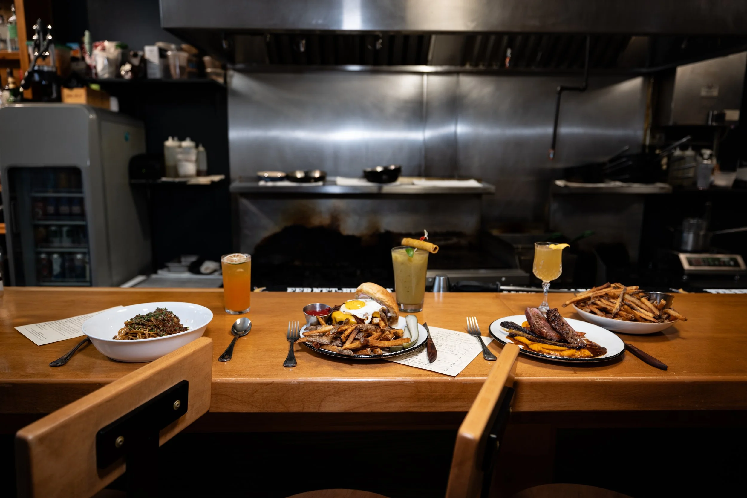 A wooden restaurant counter with various plates of food, including a bowl of pasta, a burger with fries, a steak with sauce, a plate of fries, and three drinks. The kitchen is in the background.