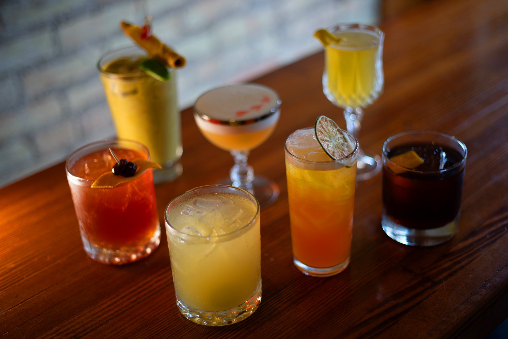A variety of colorful cocktails on a wooden table, with some garnished with fruit slices and herbs.